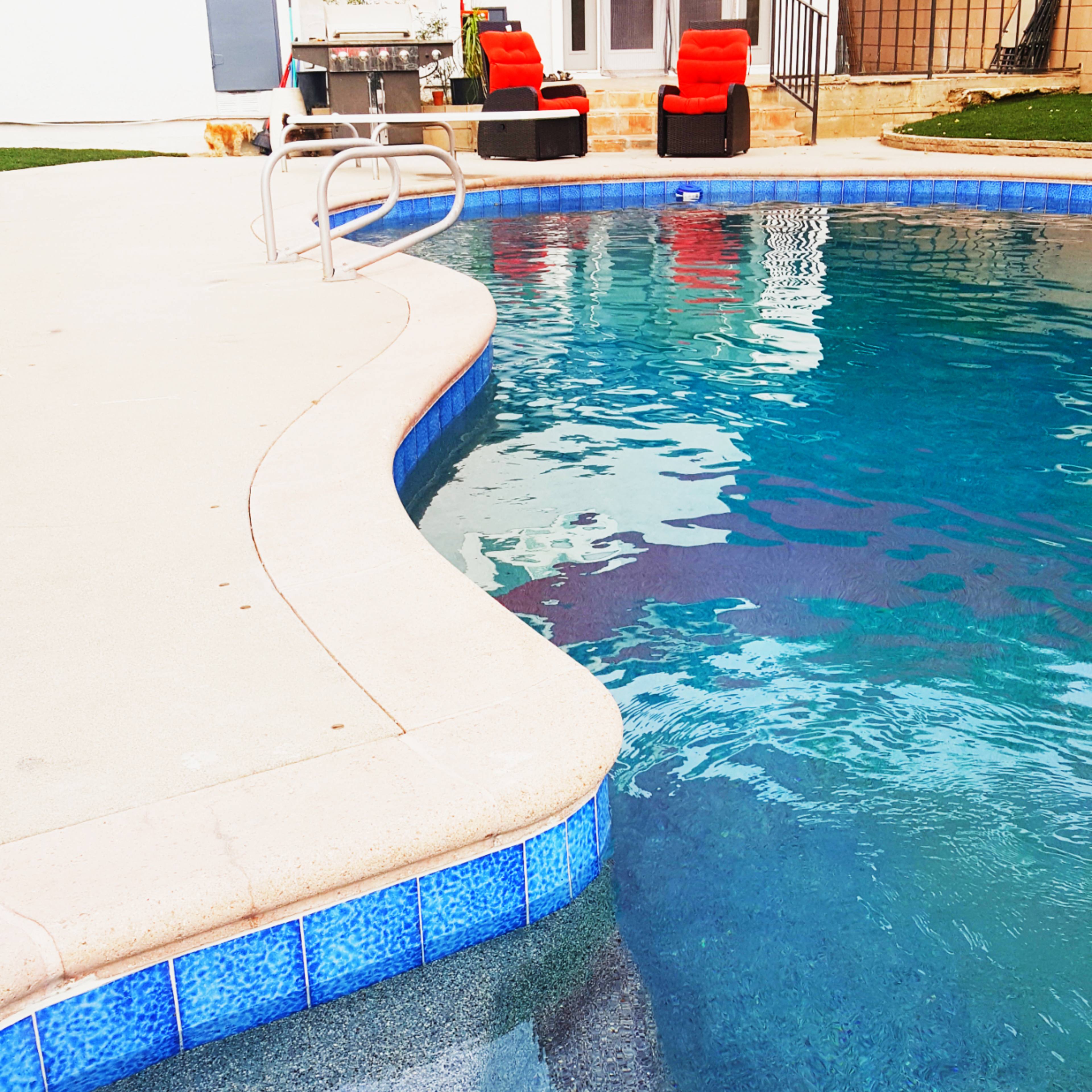 A swimming pool with a curved edge and a red shade structure over lounge chairs, located near a white building with large glass doors.