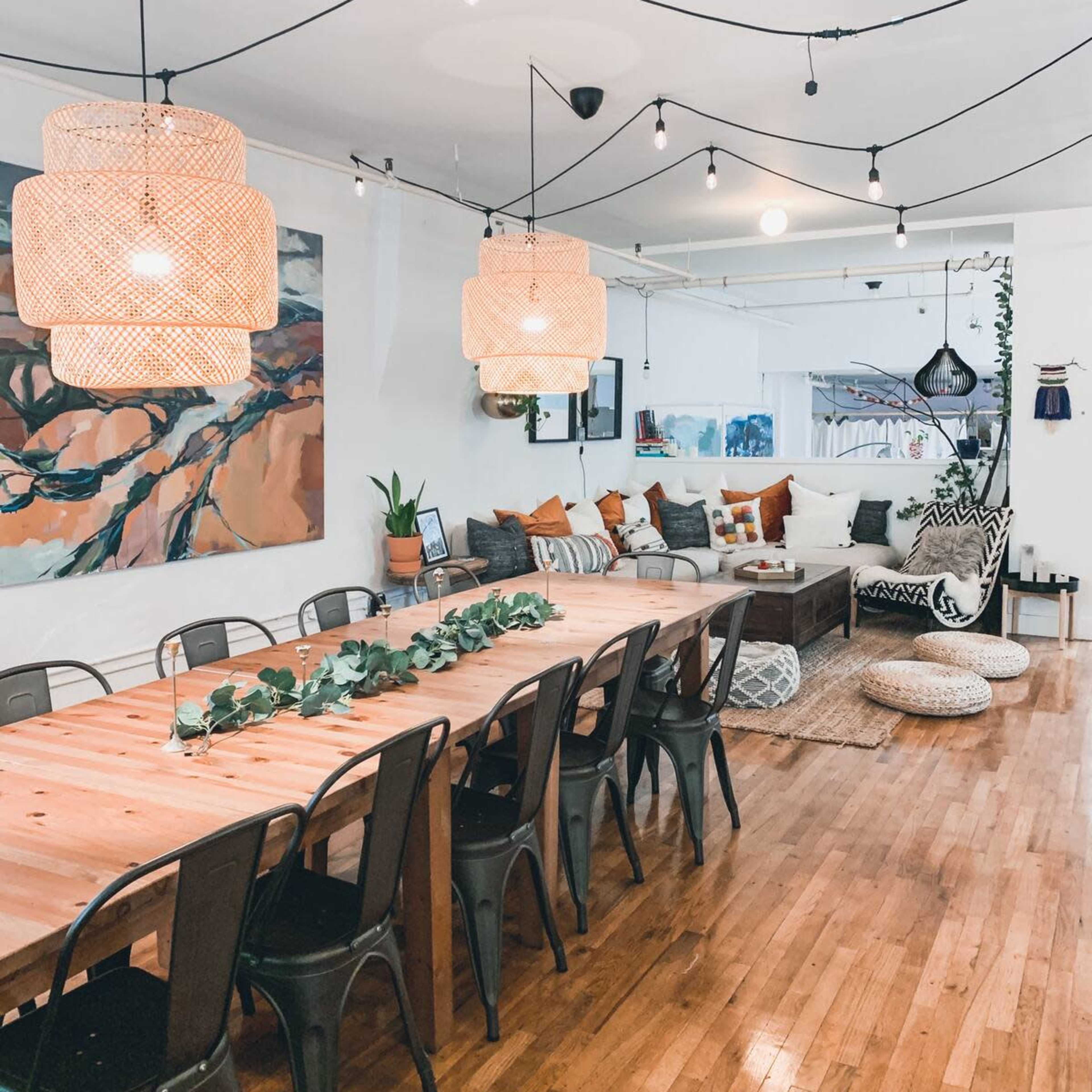 A spacious dining area with a long wooden table surrounded by metal chairs, illuminated by pendant lights and decorated with plants and artwork.