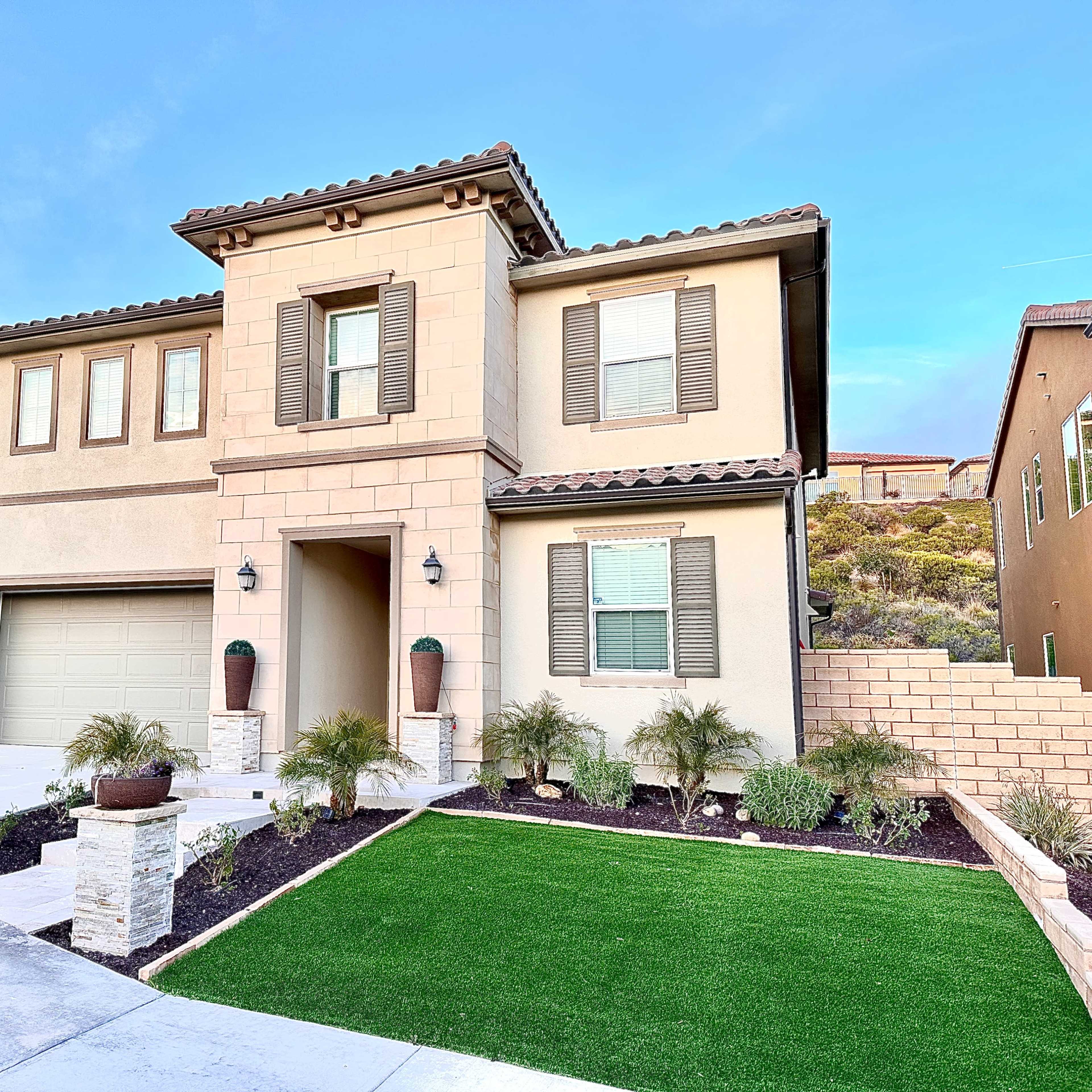 A two-story house with a stone facade, a two-car garage, and well-maintained landscaping featuring potted plants and artificial grass.
