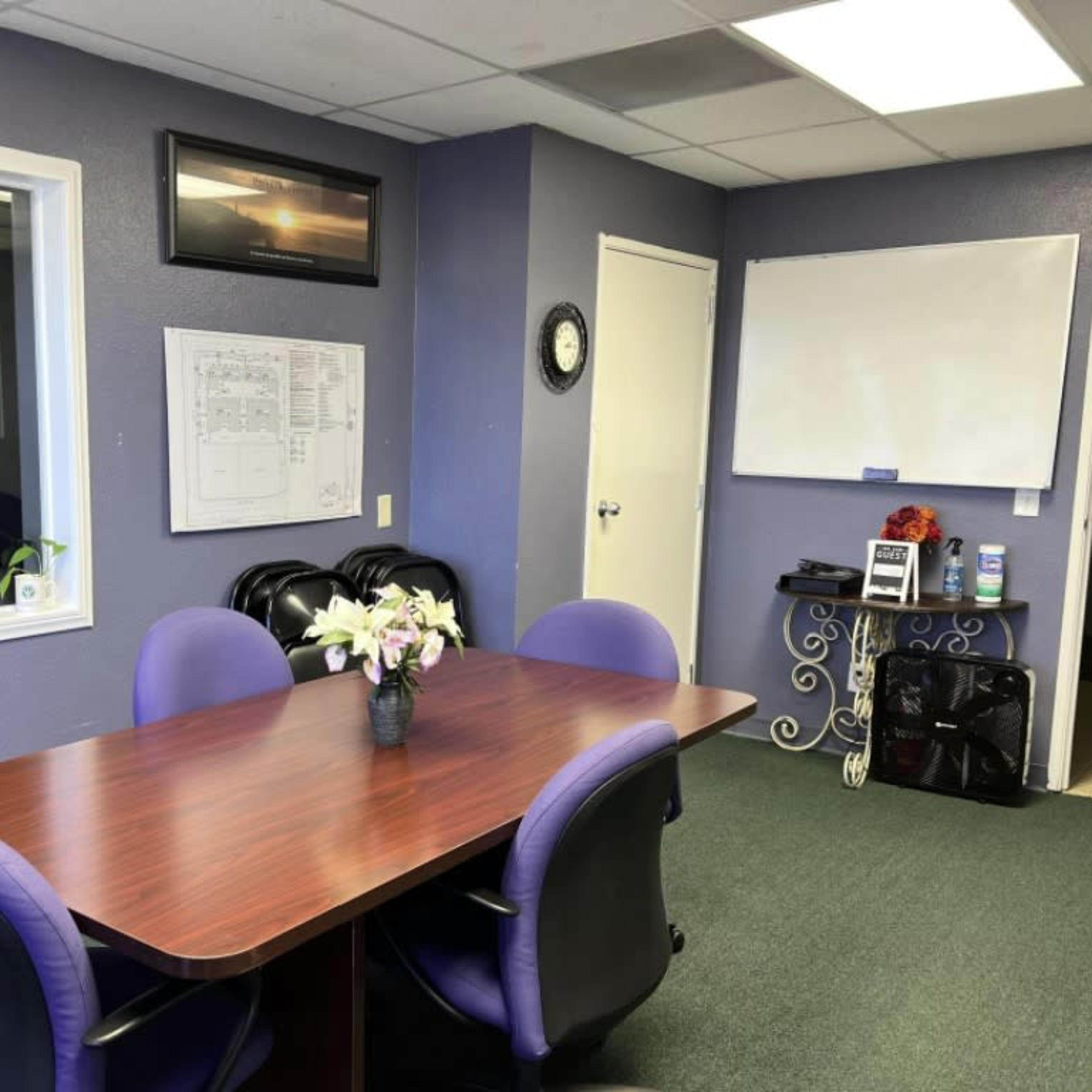 A conference room with a brown table surrounded by black and purple chairs, a whiteboard, and a window overlooking another room.