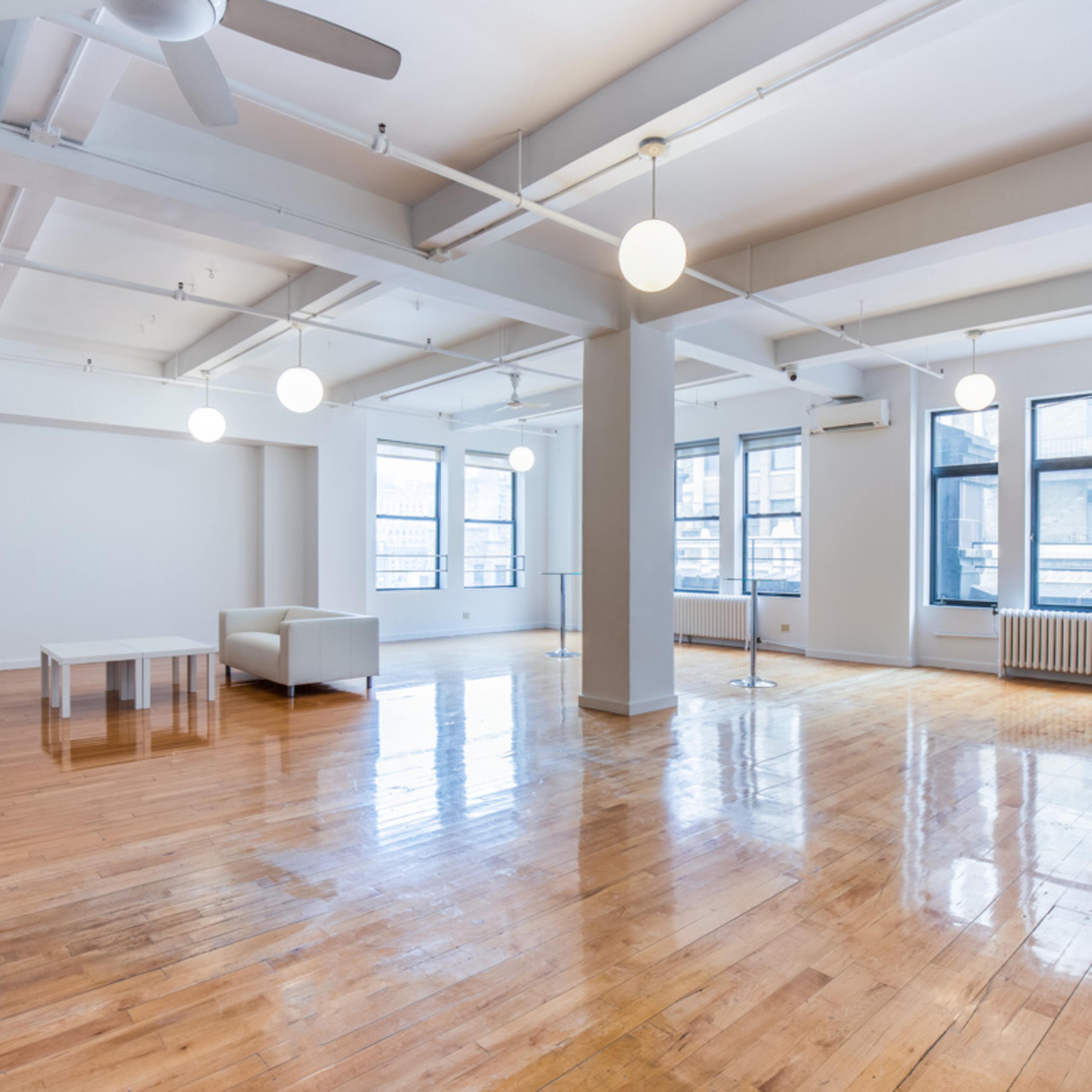 A spacious, empty room with hardwood floors, large windows, and minimalist furniture.