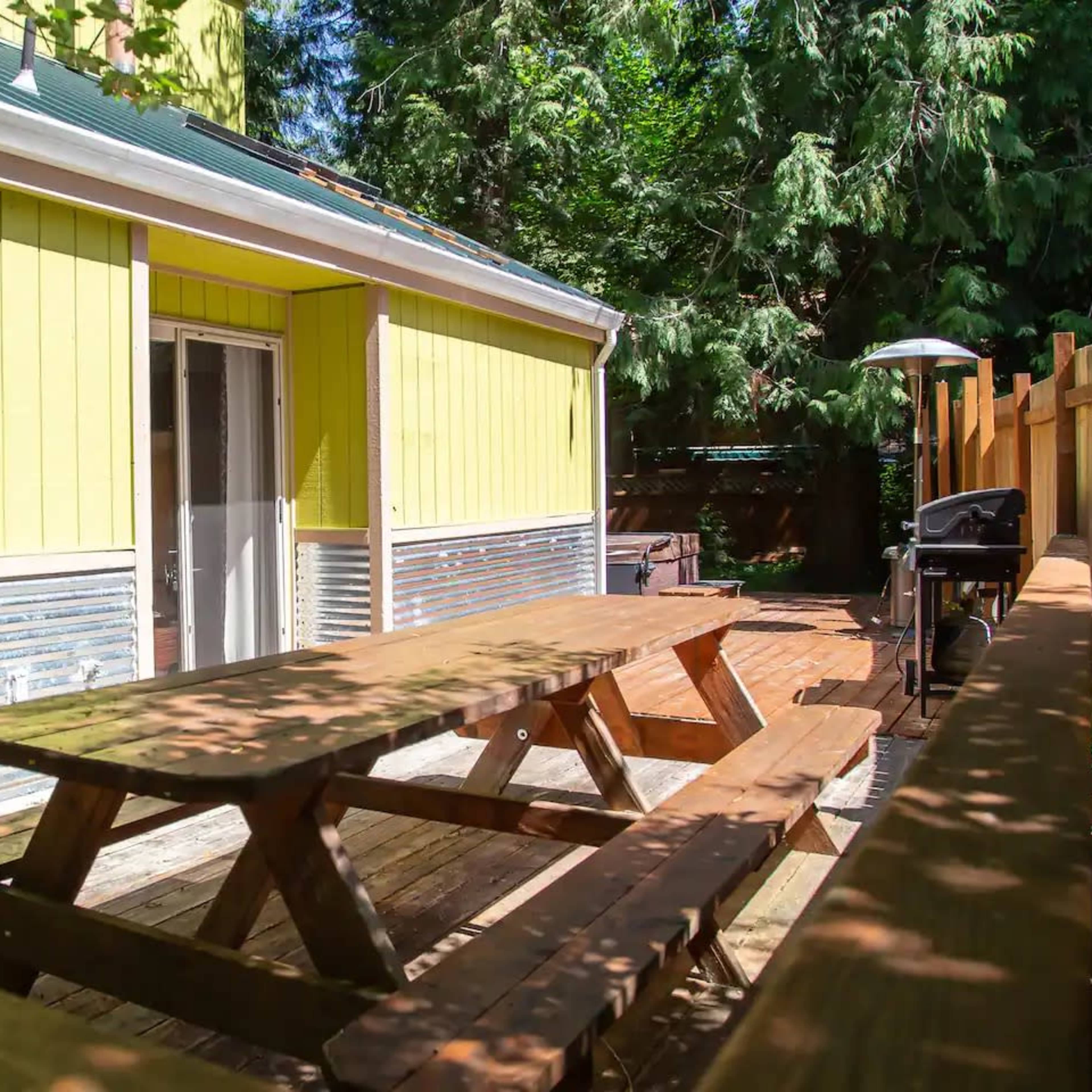 A yellow house features a wooden deck with multiple picnic tables and surrounding greenery.