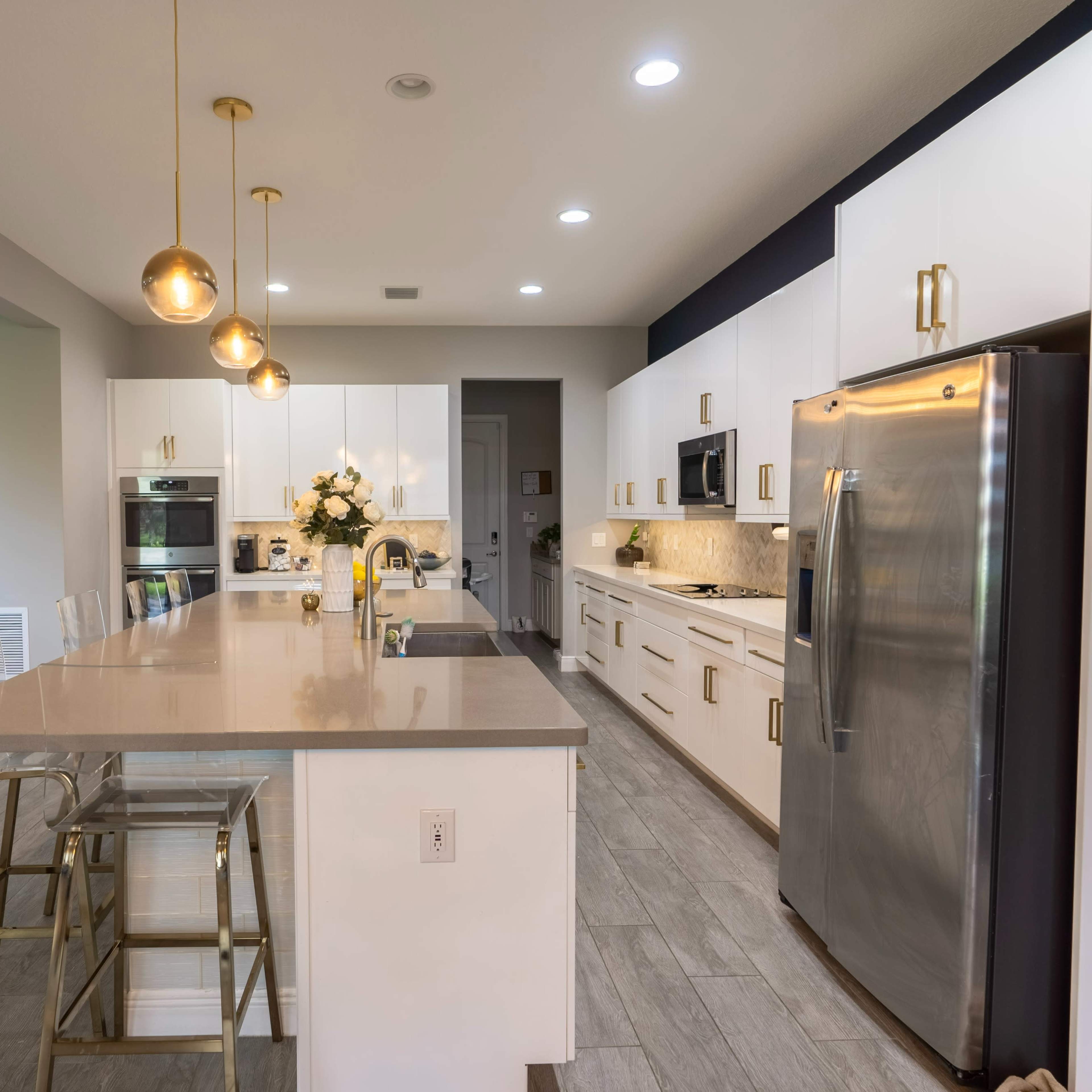 A modern kitchen with white cabinetry, a stainless steel refrigerator, a large island with bar stools, and pendant lighting.