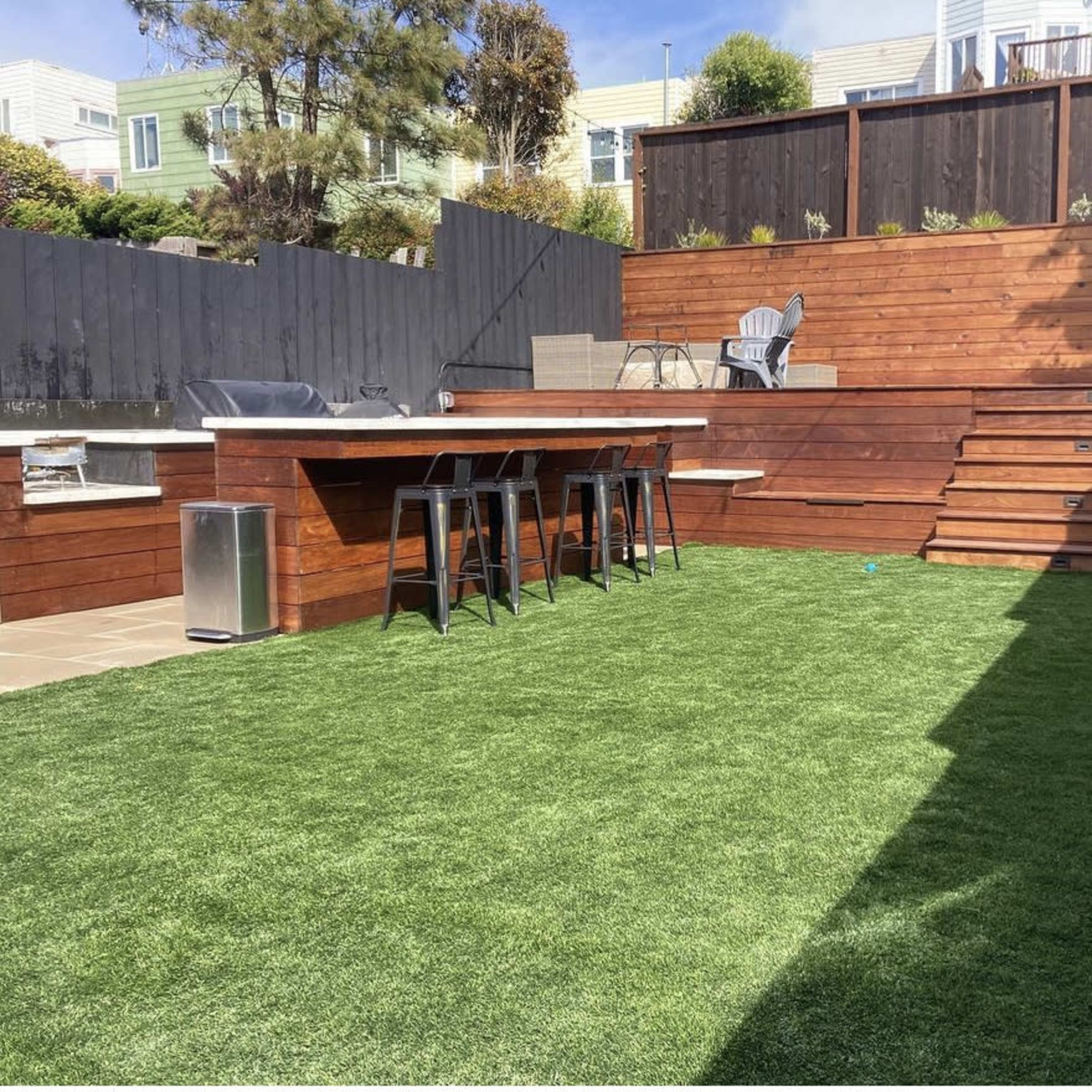 A backyard with artificial grass, wooden seating areas, a bar with stools, and steps leading to a higher level, surrounded by wooden fencing and landscaping.