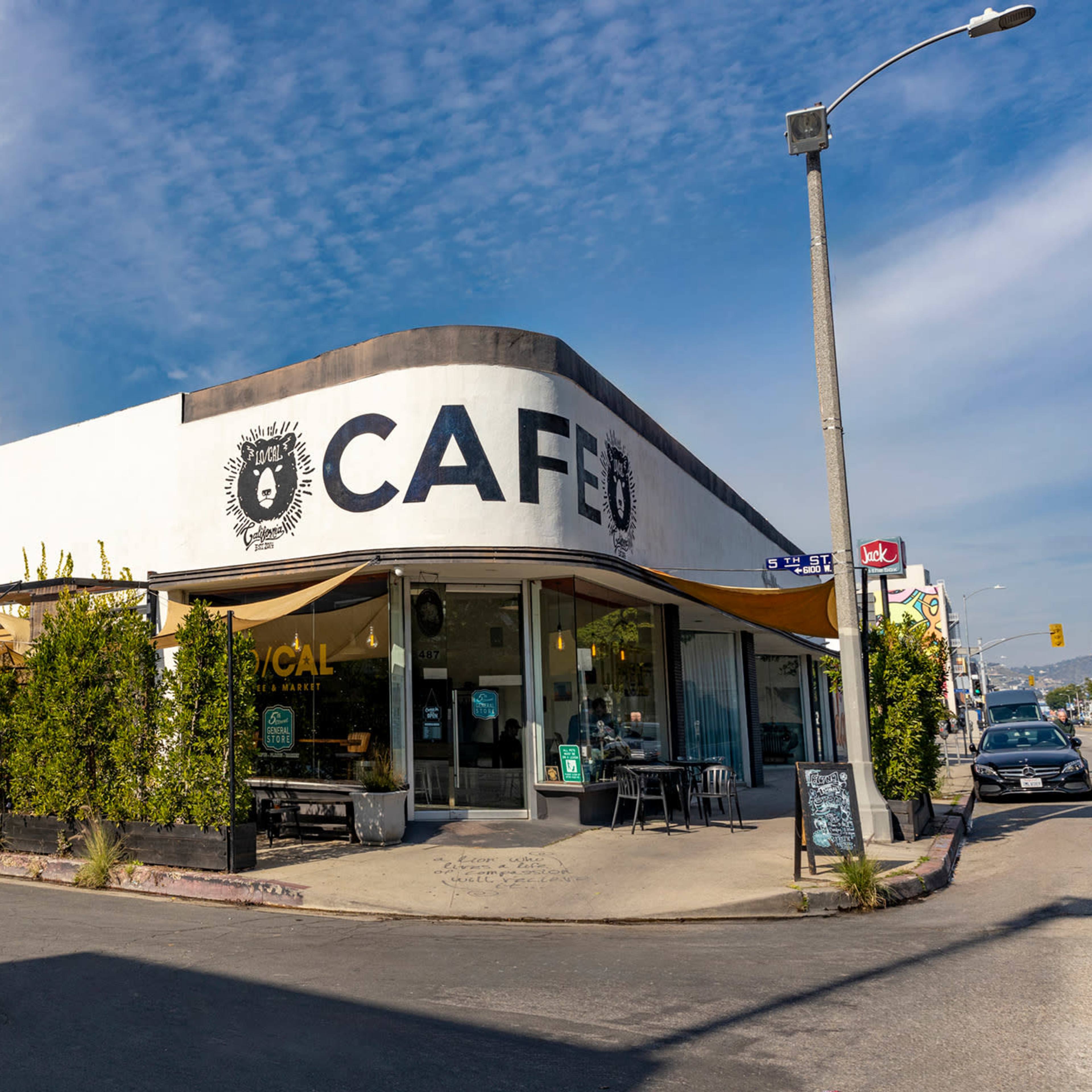 A corner cafe features a white exterior with large black lettering and outdoor seating surrounded by greenery.