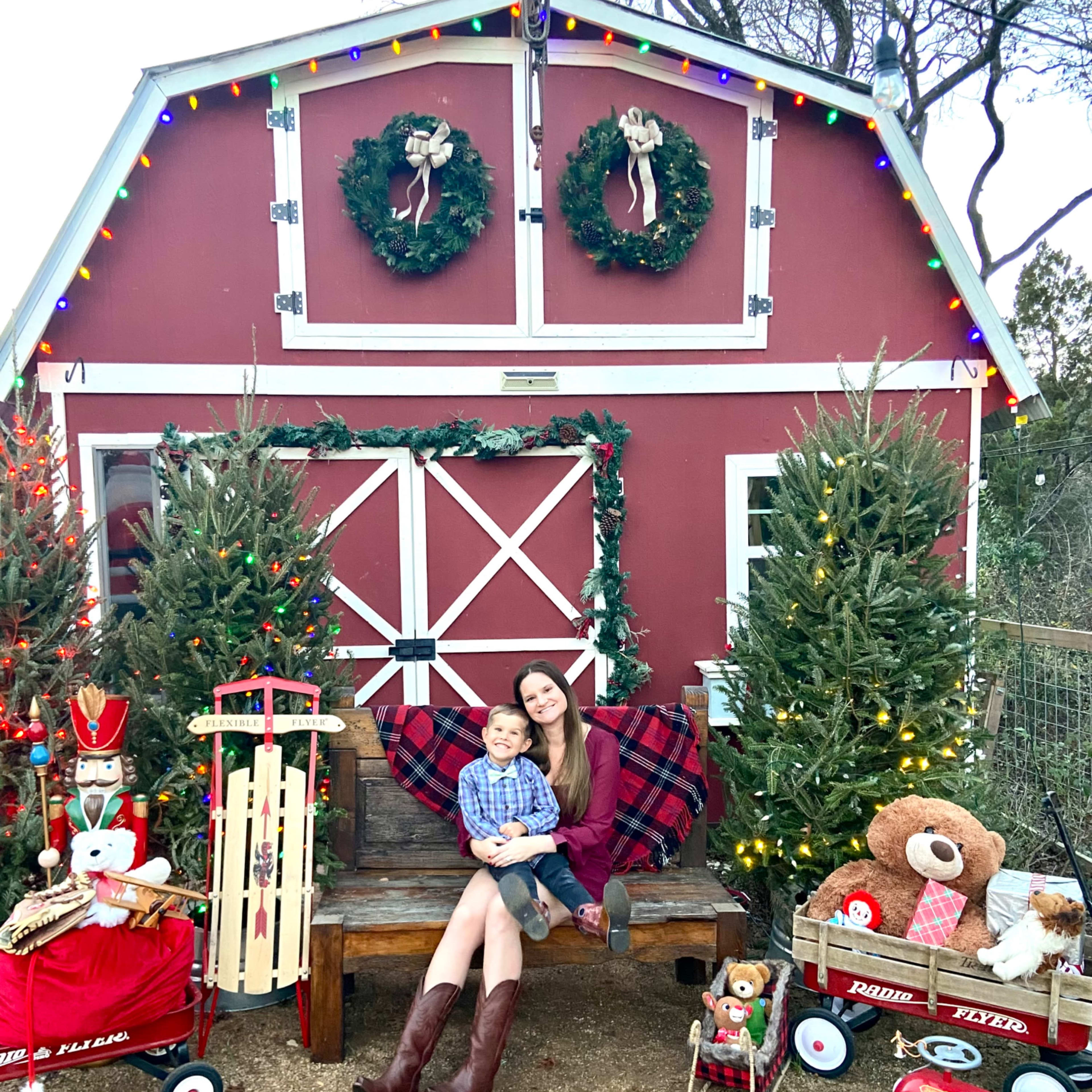 A woman and a child sit on a bench next to a festive red barn decorated with Christmas wreaths and surrounded by trees adorned with lights.