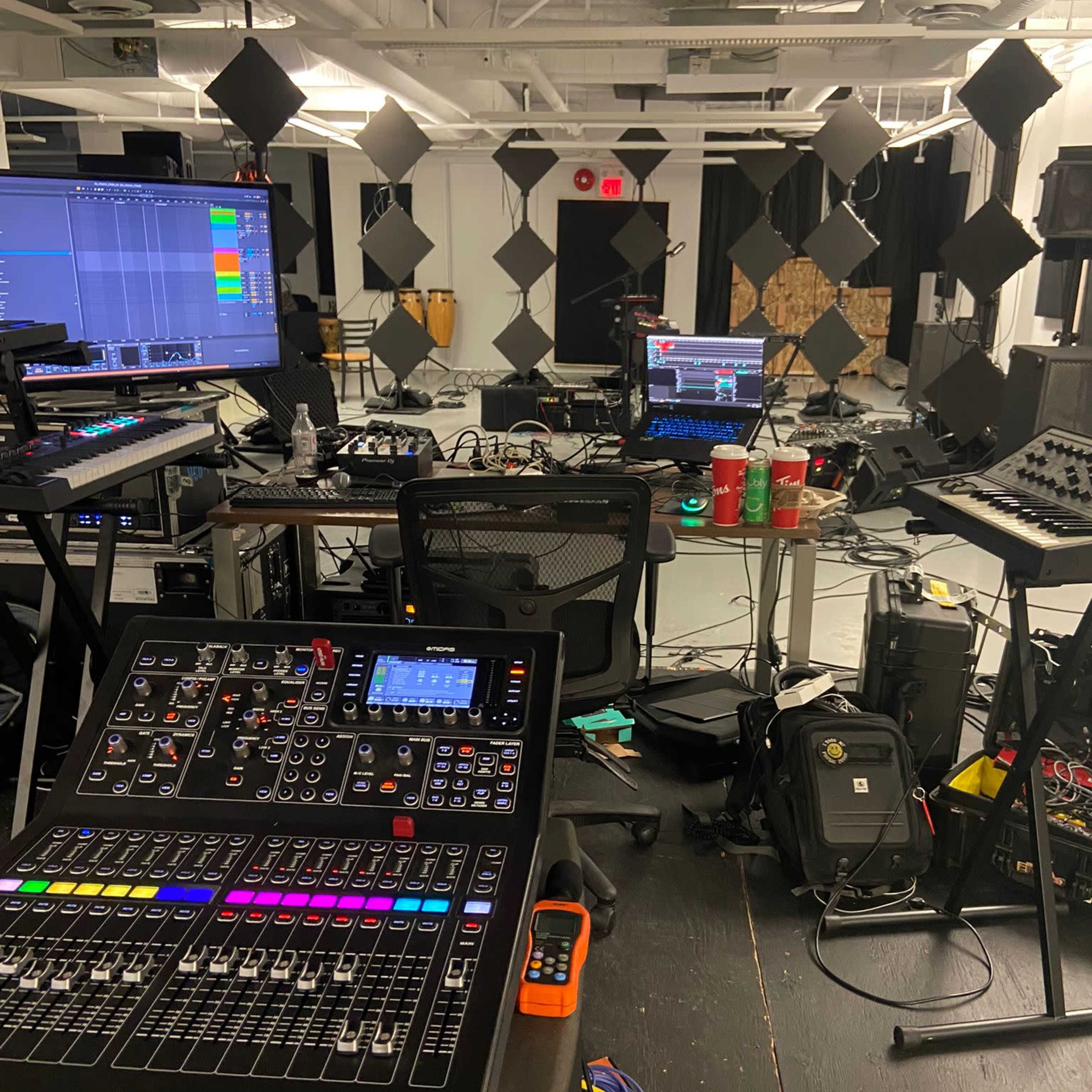 A music studio filled with various electronic equipment, including mixers and computers, alongside scattered cables and cups on a table.