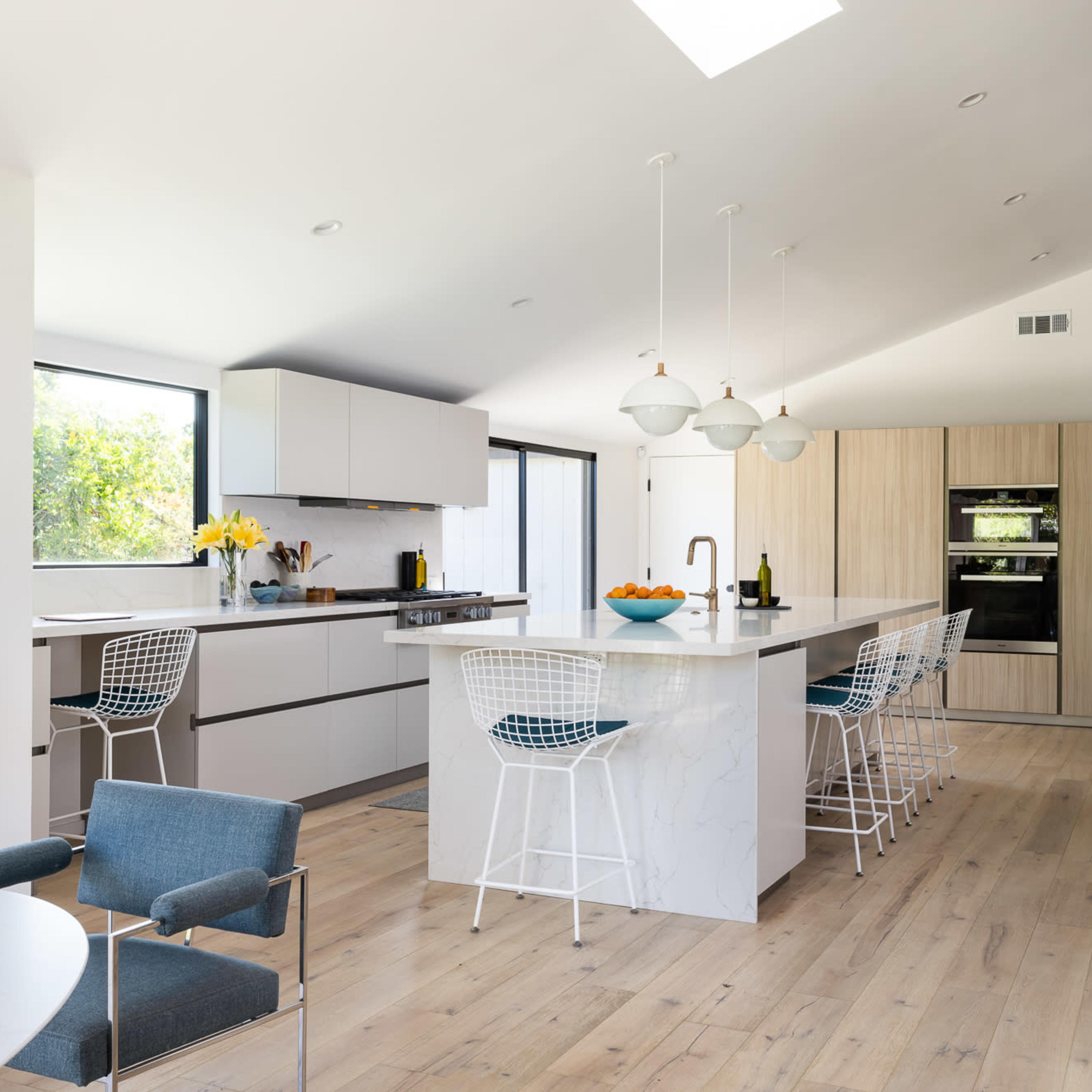 A modern kitchen features a central island with bar stools, large windows, and a combination of light-colored cabinetry and wood accents.