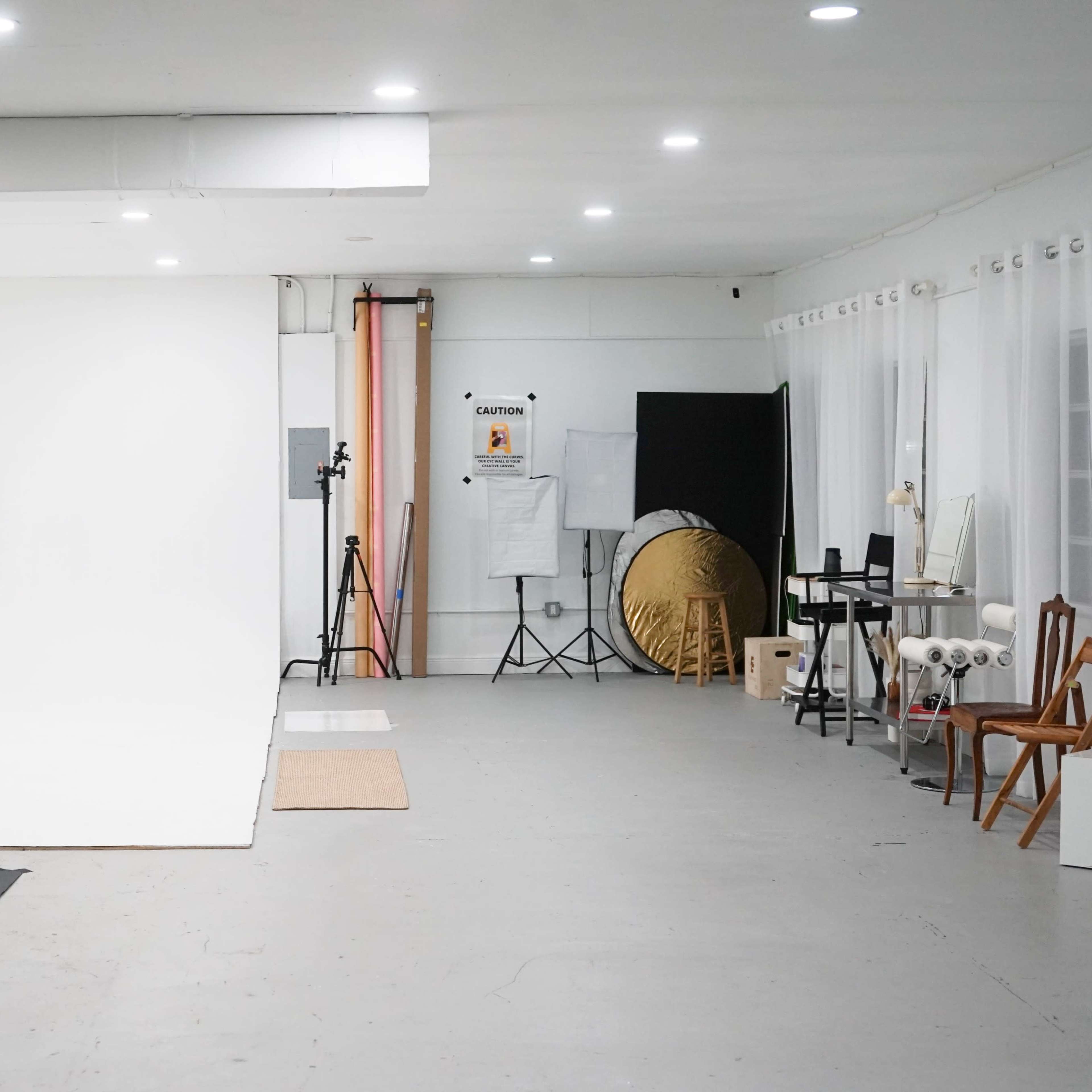 A photography studio with a blank backdrop, various lighting equipment, and furniture arranged along the walls.