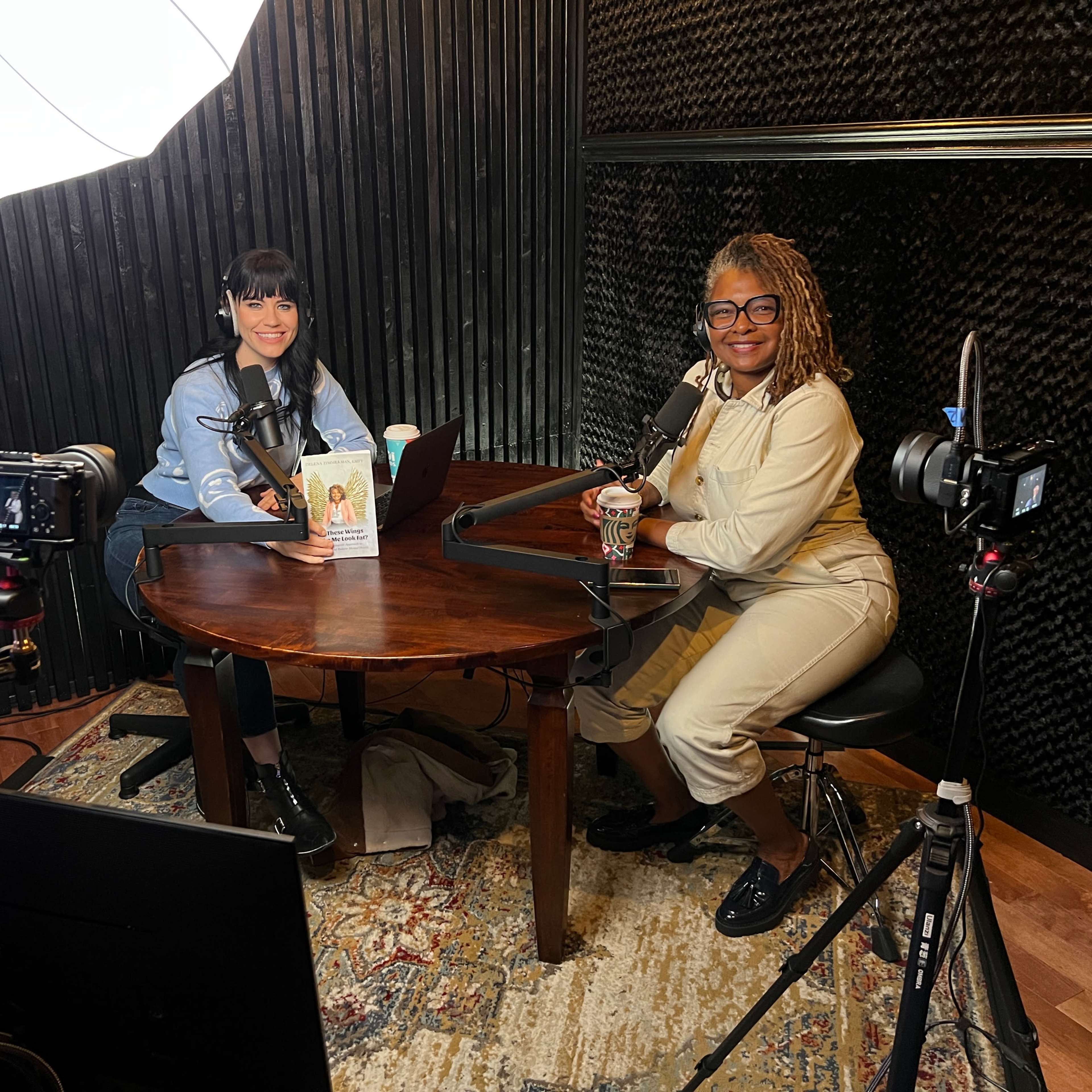 Two women are seated at a table in a podcast studio, equipped with microphones and camera equipment, with a large softbox light overhead.