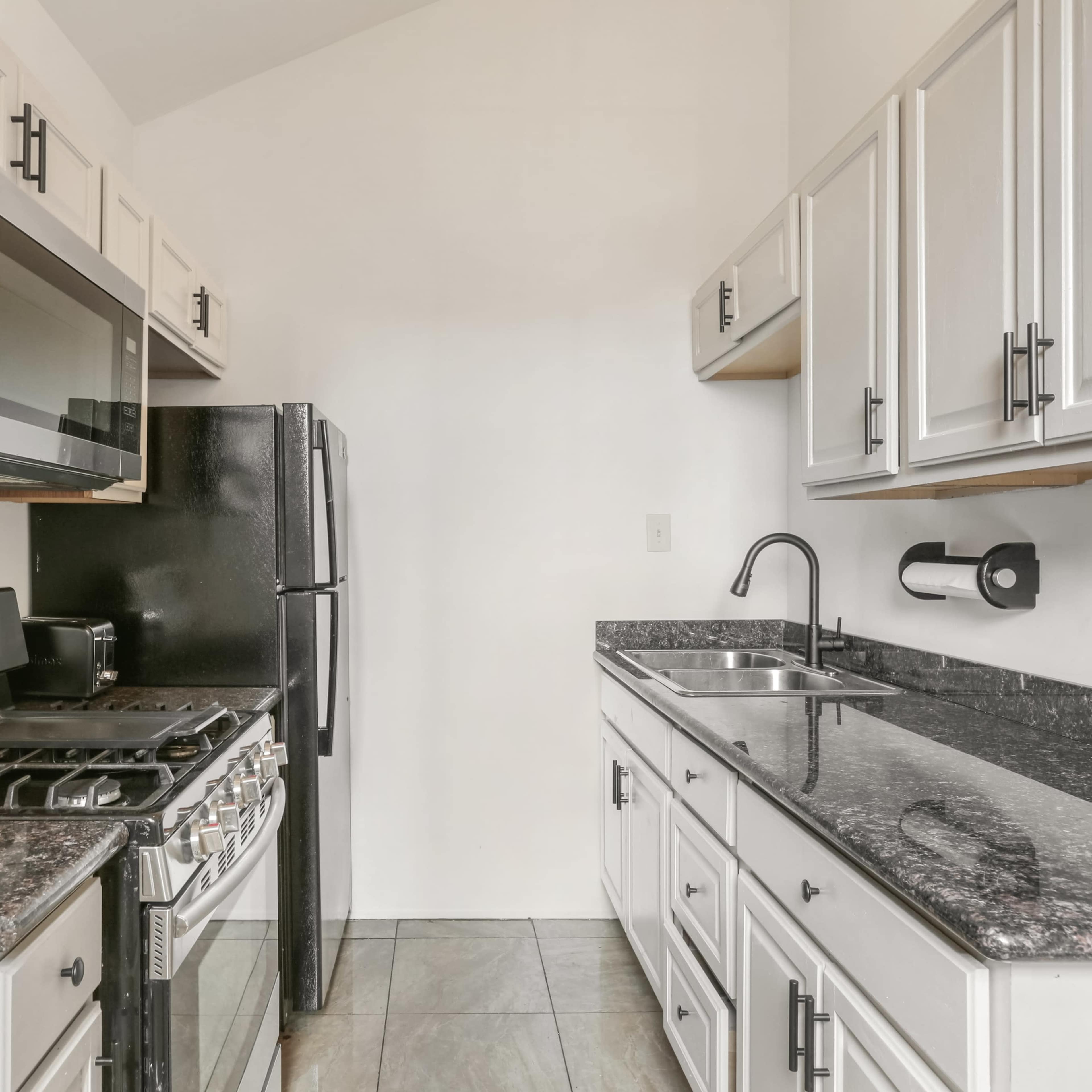 A compact kitchen features white cabinetry, a black refrigerator, a stainless steel gas stove, and a sink set against dark granite countertops.