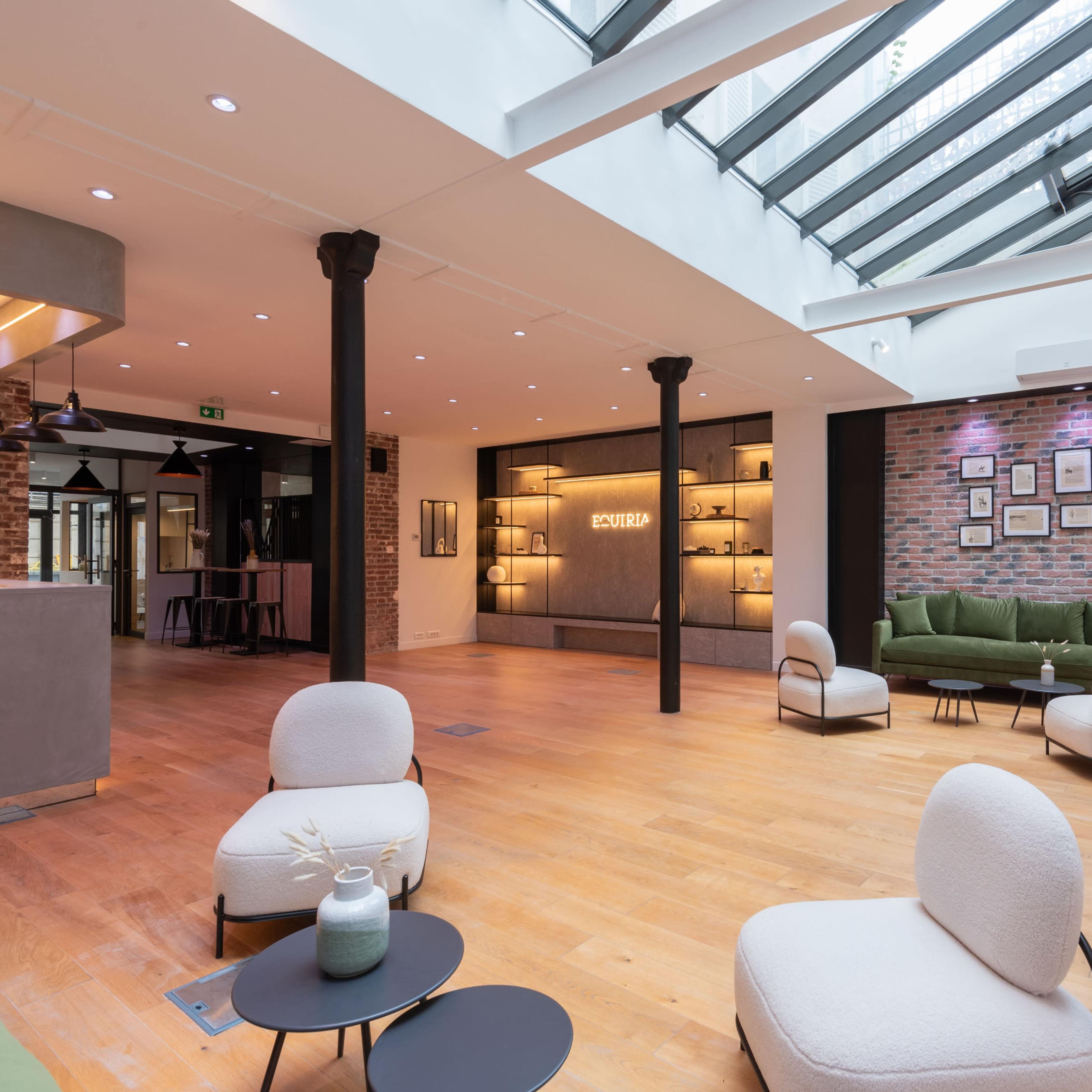 A modern interior space with wooden flooring, featuring green sofas, minimalist chairs, and a glass ceiling that allows natural light to flood in.