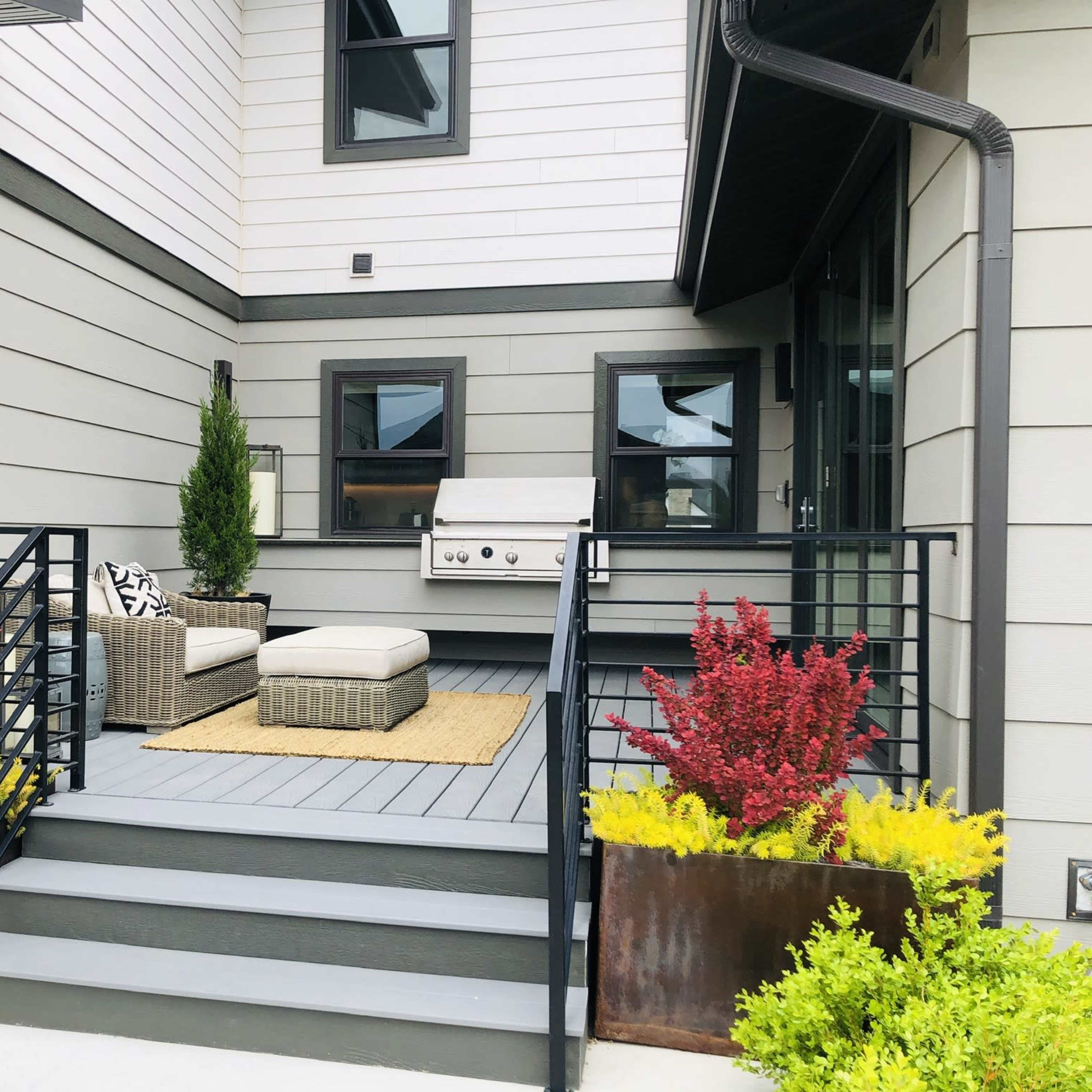 A modern porch features a grill, seating area, and planters with colorful plants on either side.