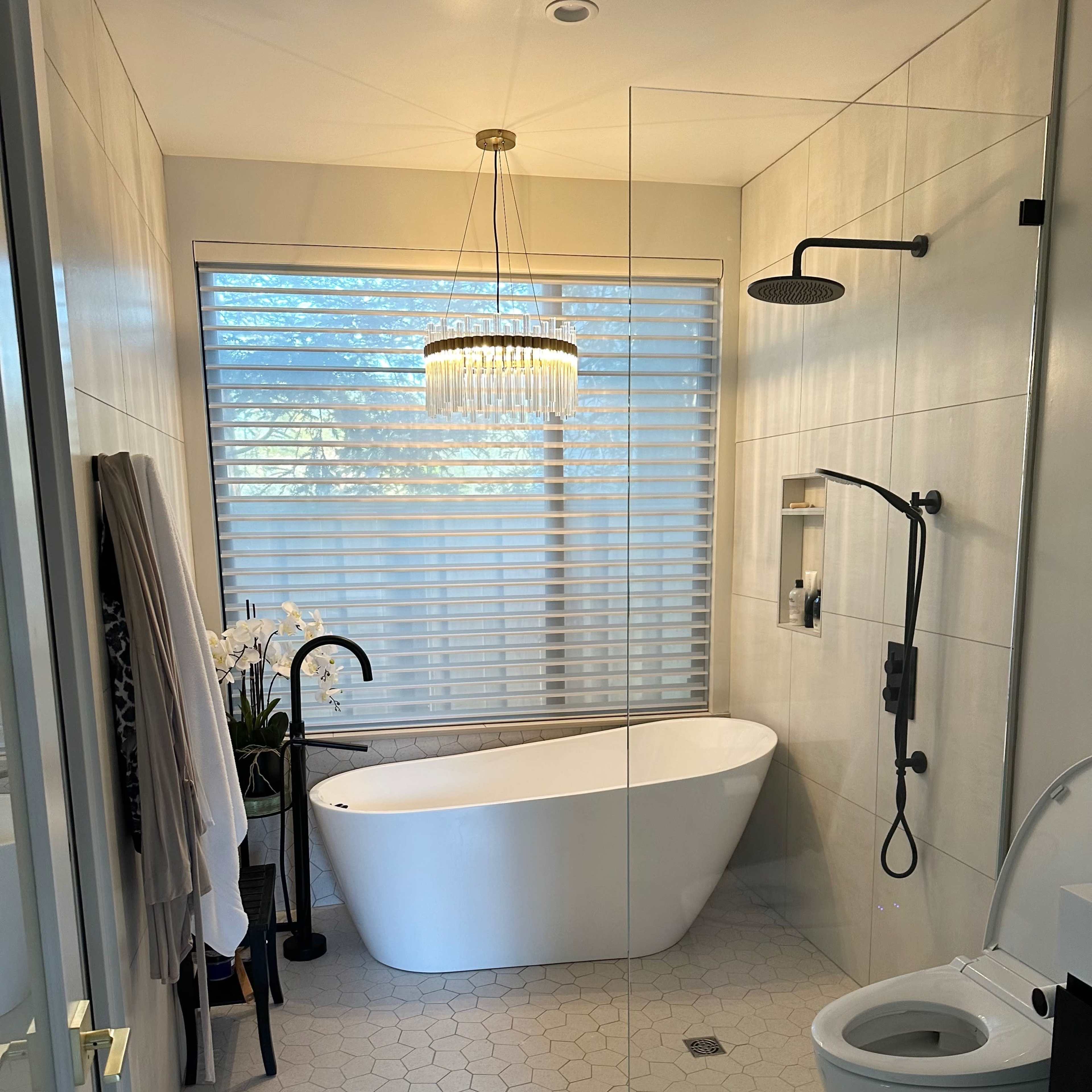 The bathroom features a freestanding tub, a glass shower enclosure, a modern toilet, and a stylish chandelier above, with large windows allowing natural light to filter through the blinds.