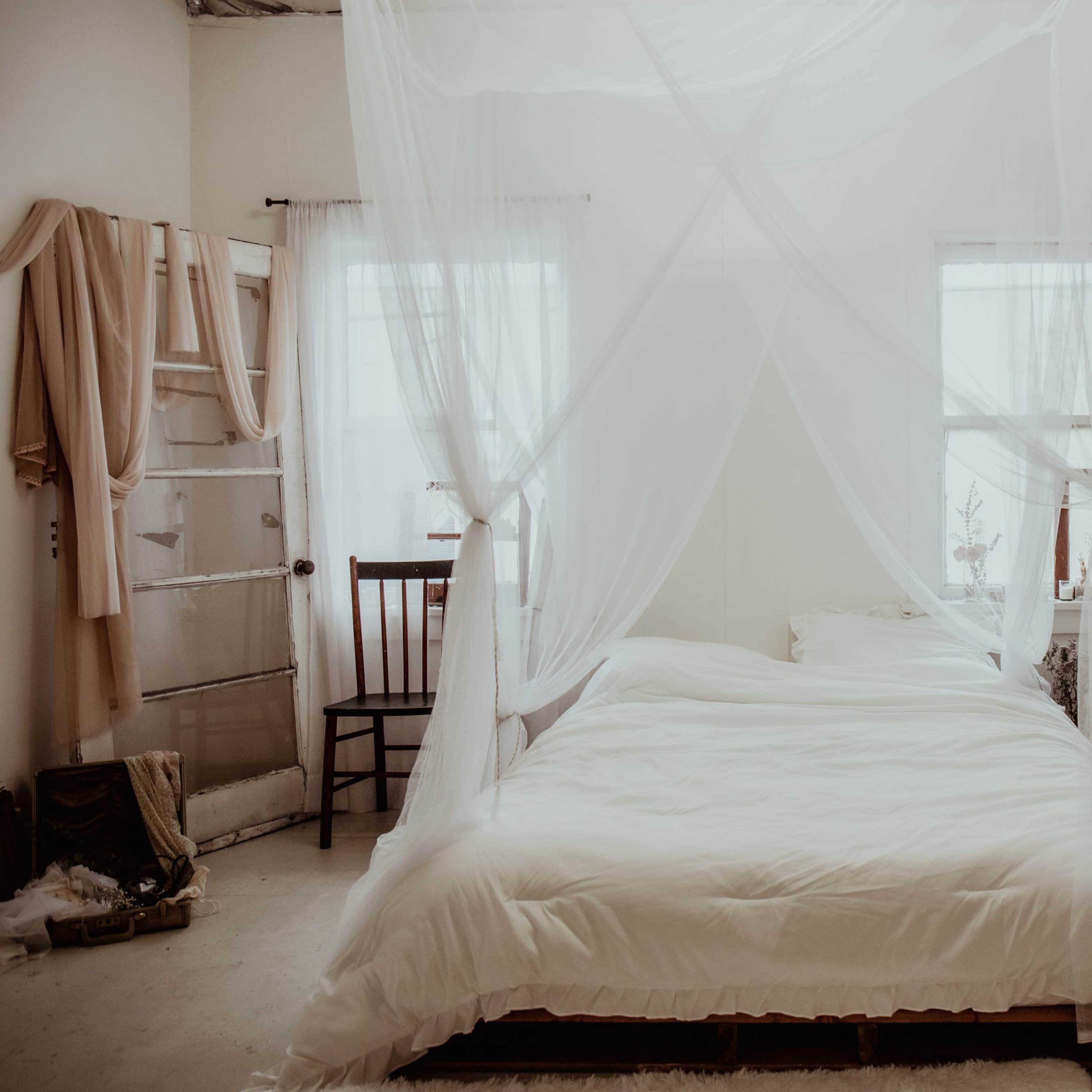 A minimalist bedroom with a large bed draped in sheer white fabric, two windows, and a wooden chair.