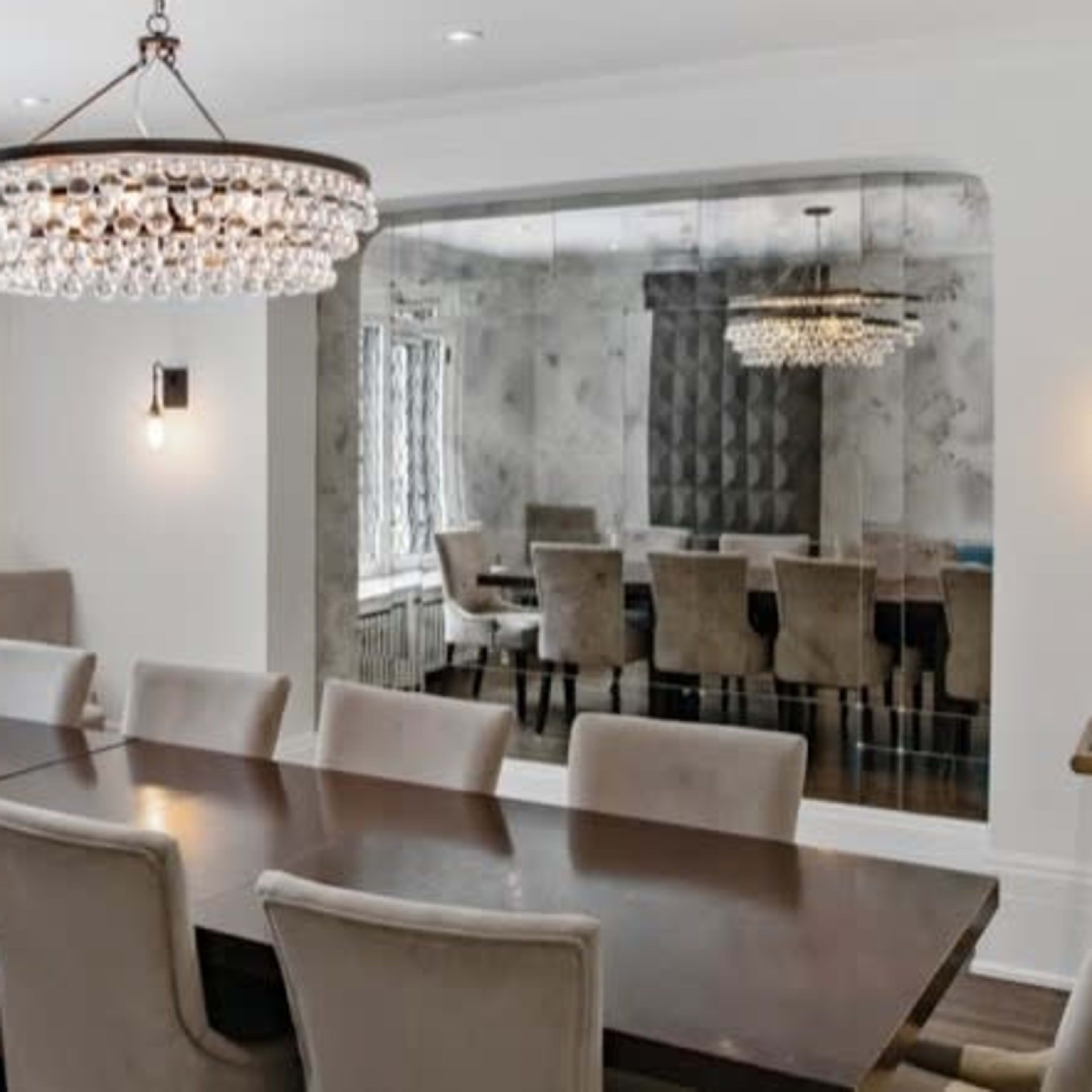 A spacious dining room with a large wooden table surrounded by upholstered chairs, featuring a chandelier and a mirrored wall.