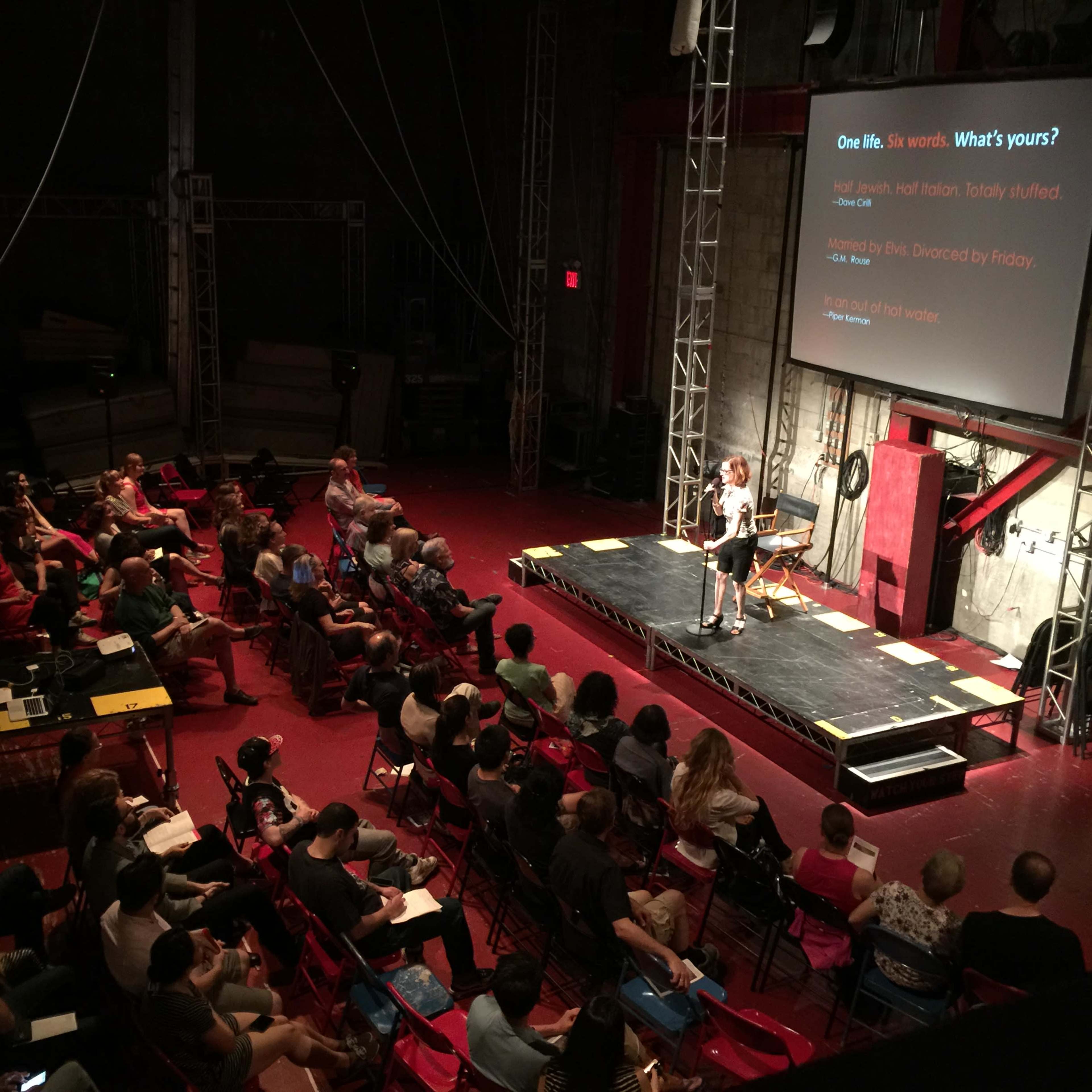 A speaker presents on stage to an audience seated in red chairs in a well-lit auditorium.