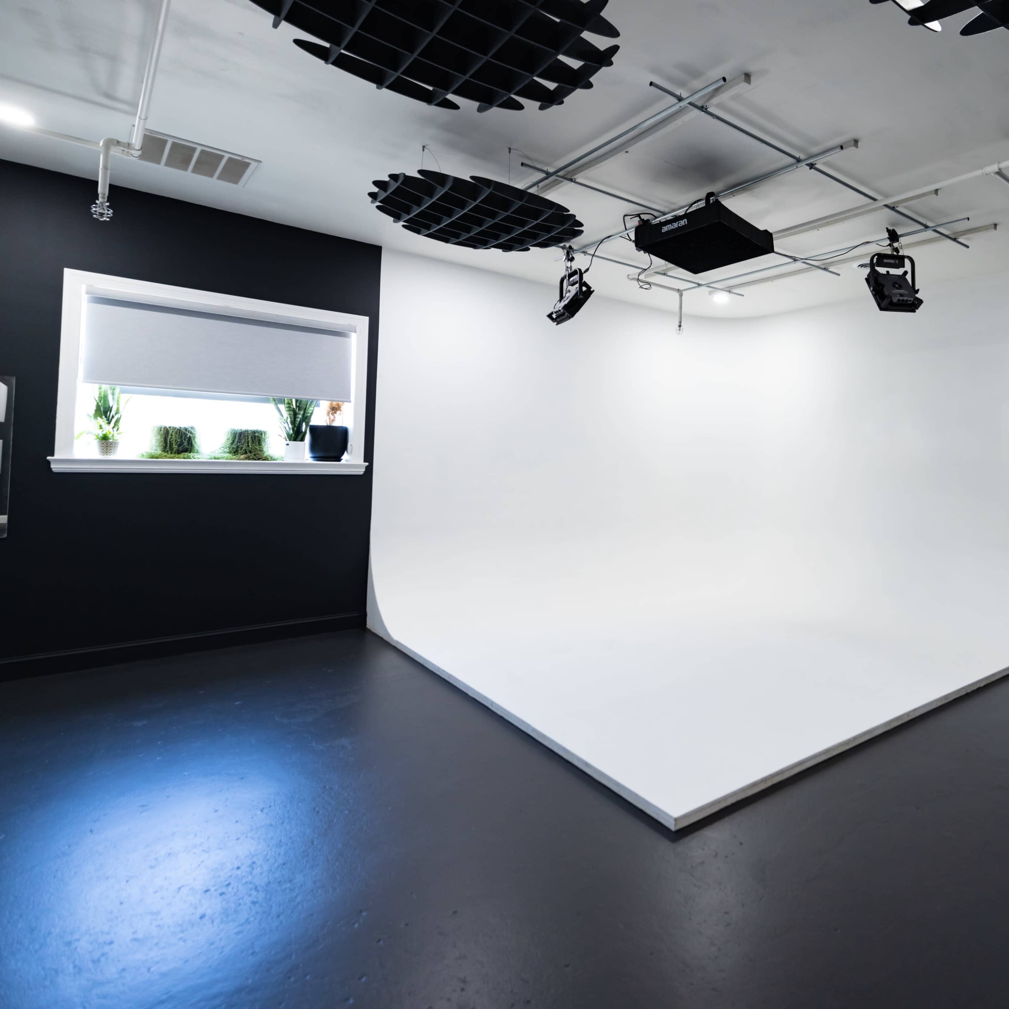 A minimalist studio space with a white cyclorama wall and dark walls, equipped with overhead lighting and a window.