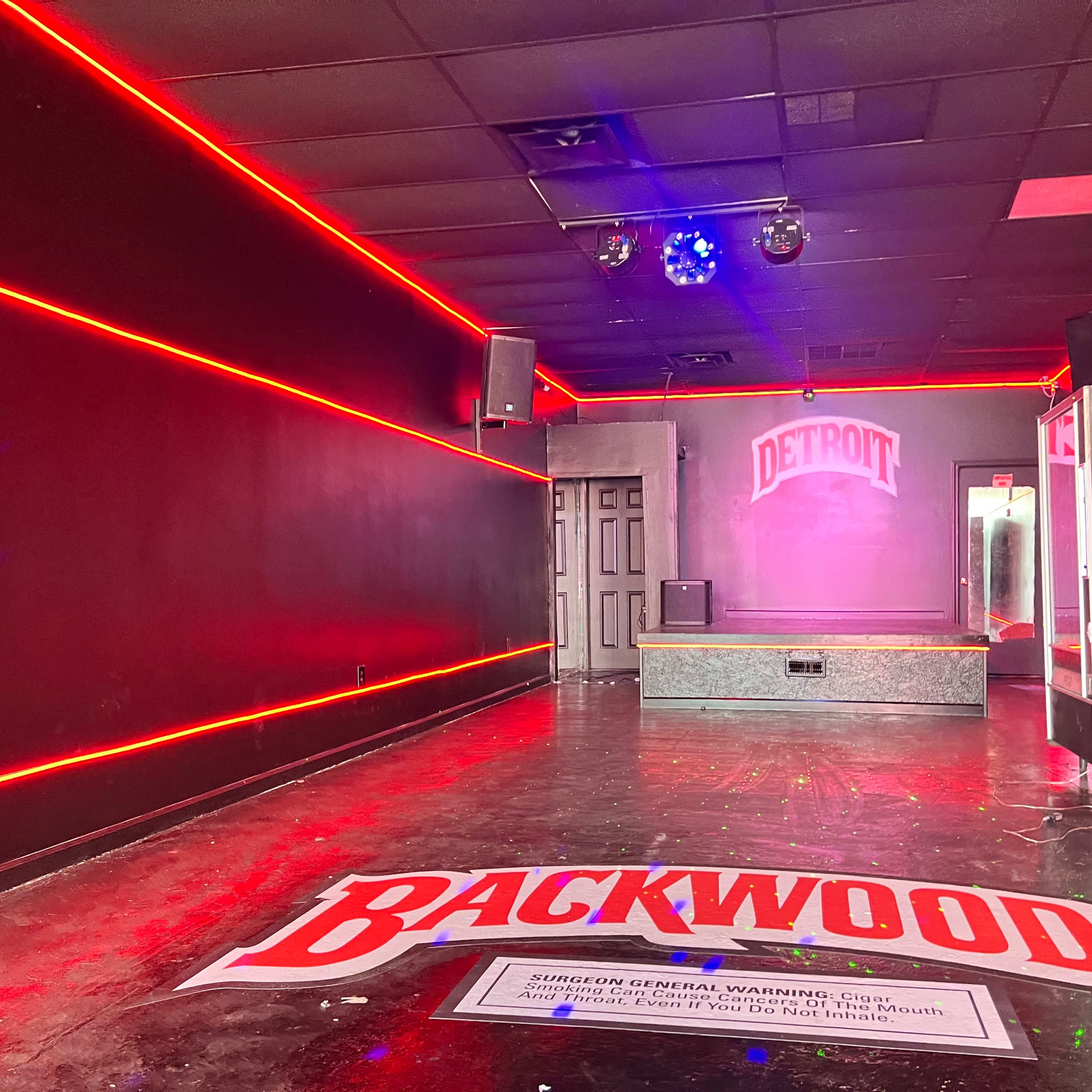 An interior space with red neon lighting, a small stage, and a large banner on the floor displaying the word "BACKWOODS."