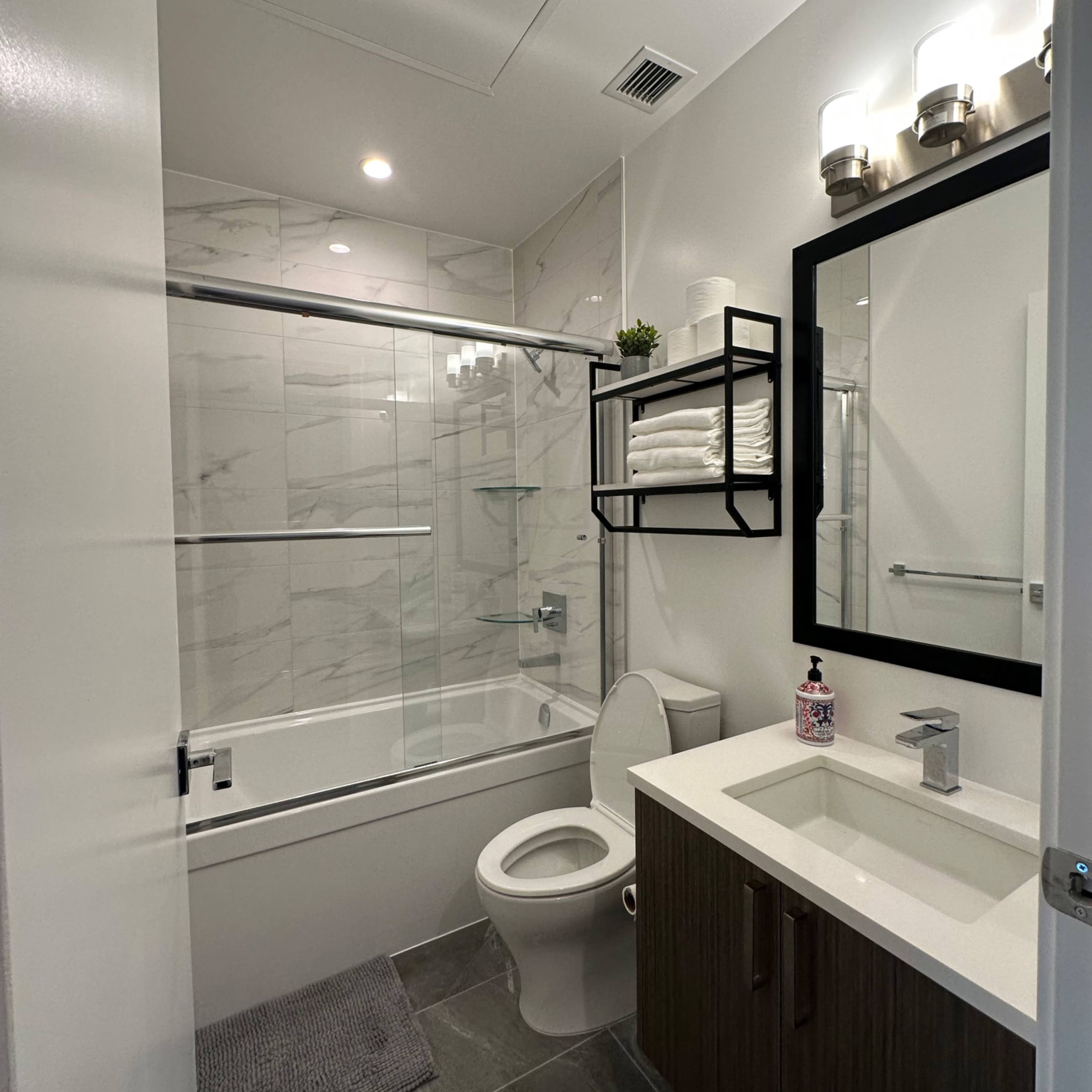 A modern bathroom featuring a bathtub with a glass shower door, a vanity with a sink, a wall-mounted shelf holding towels, and neutral-colored tile flooring.