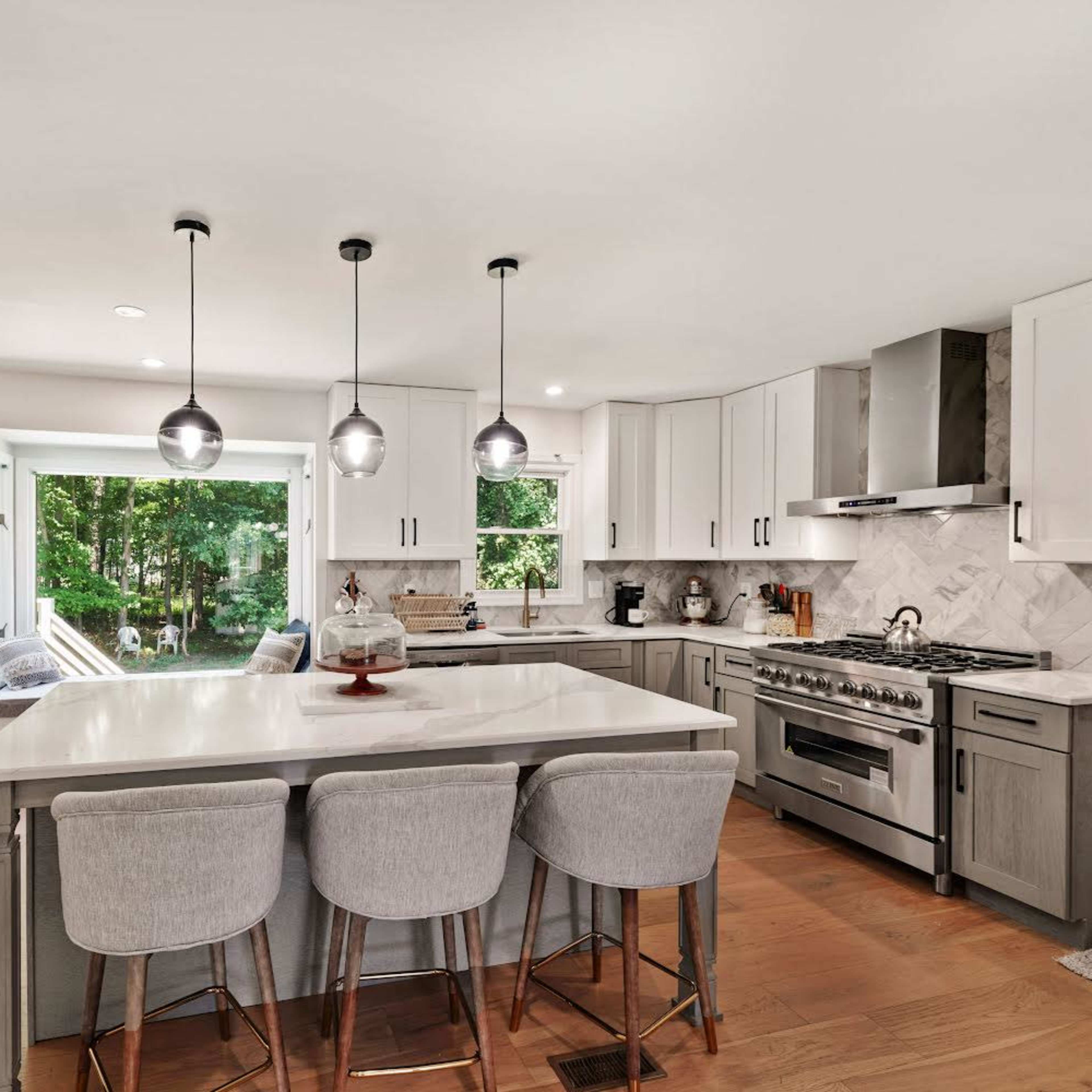 A modern kitchen features a large island with bar seating, sleek cabinetry, and large windows overlooking a wooded area.