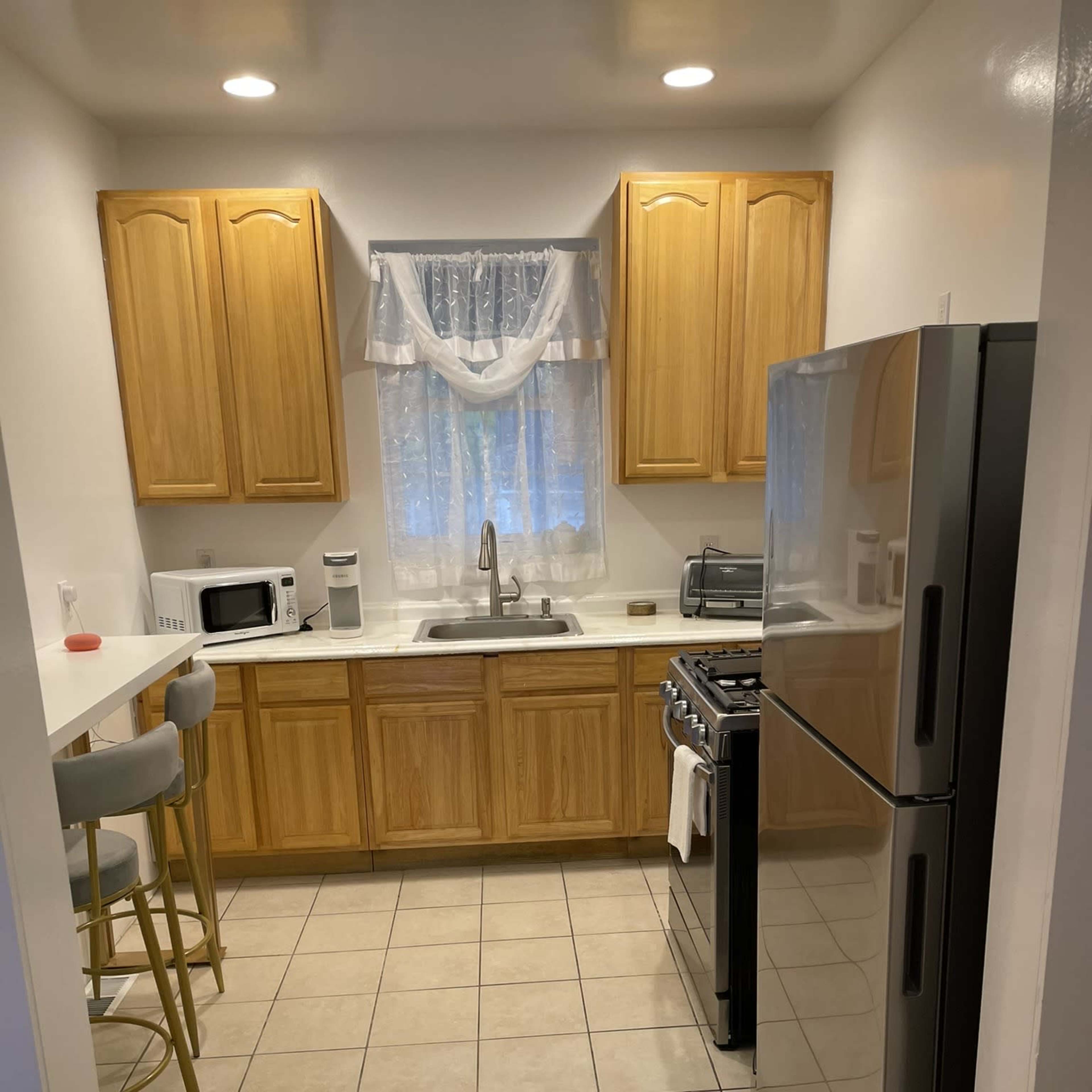 A kitchen with wooden cabinets, a sink under a window, a gas stove, a refrigerator, and a small breakfast bar with three stools.