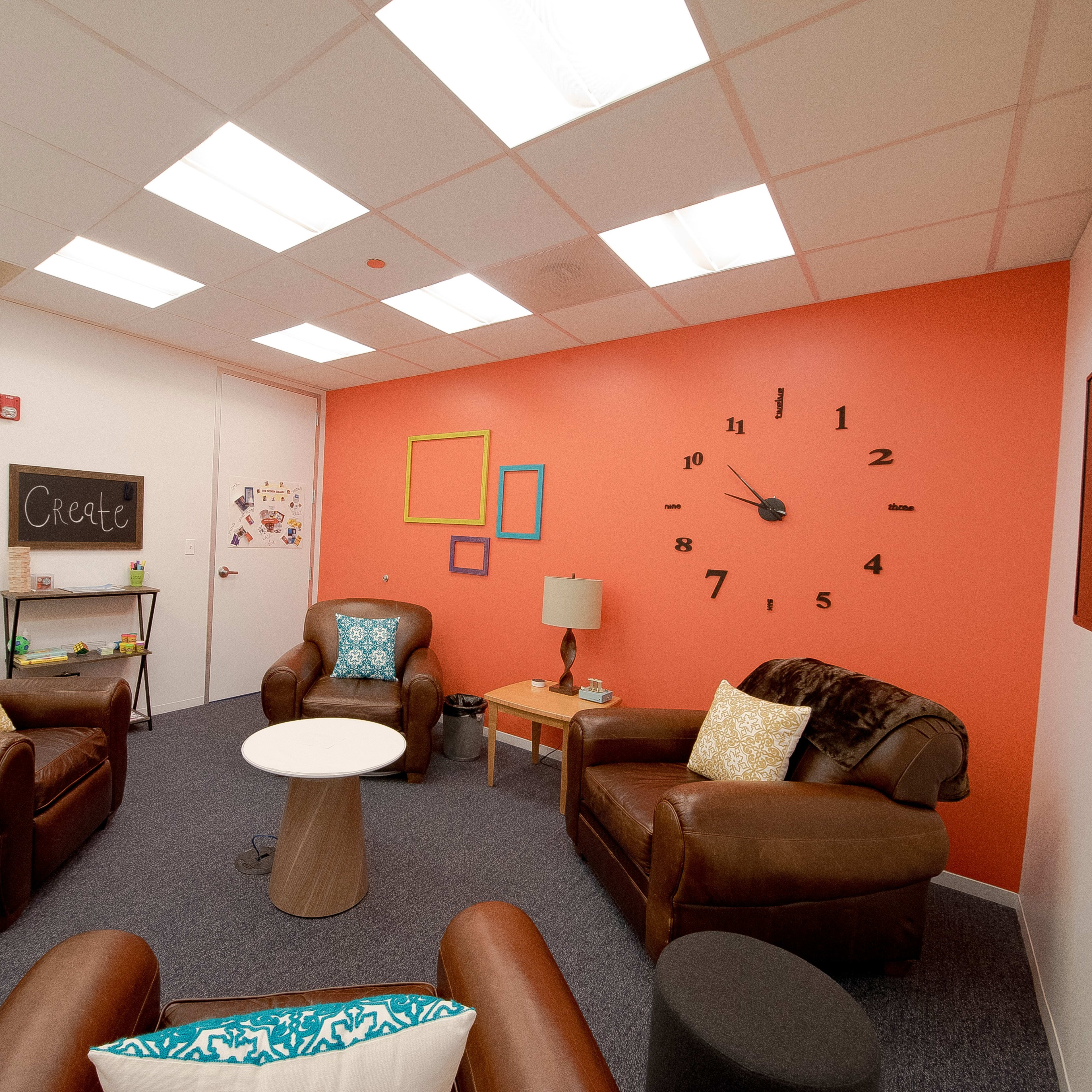 A cozy meeting room with leather chairs, a round table, a wall clock, and colorful framed decorations.