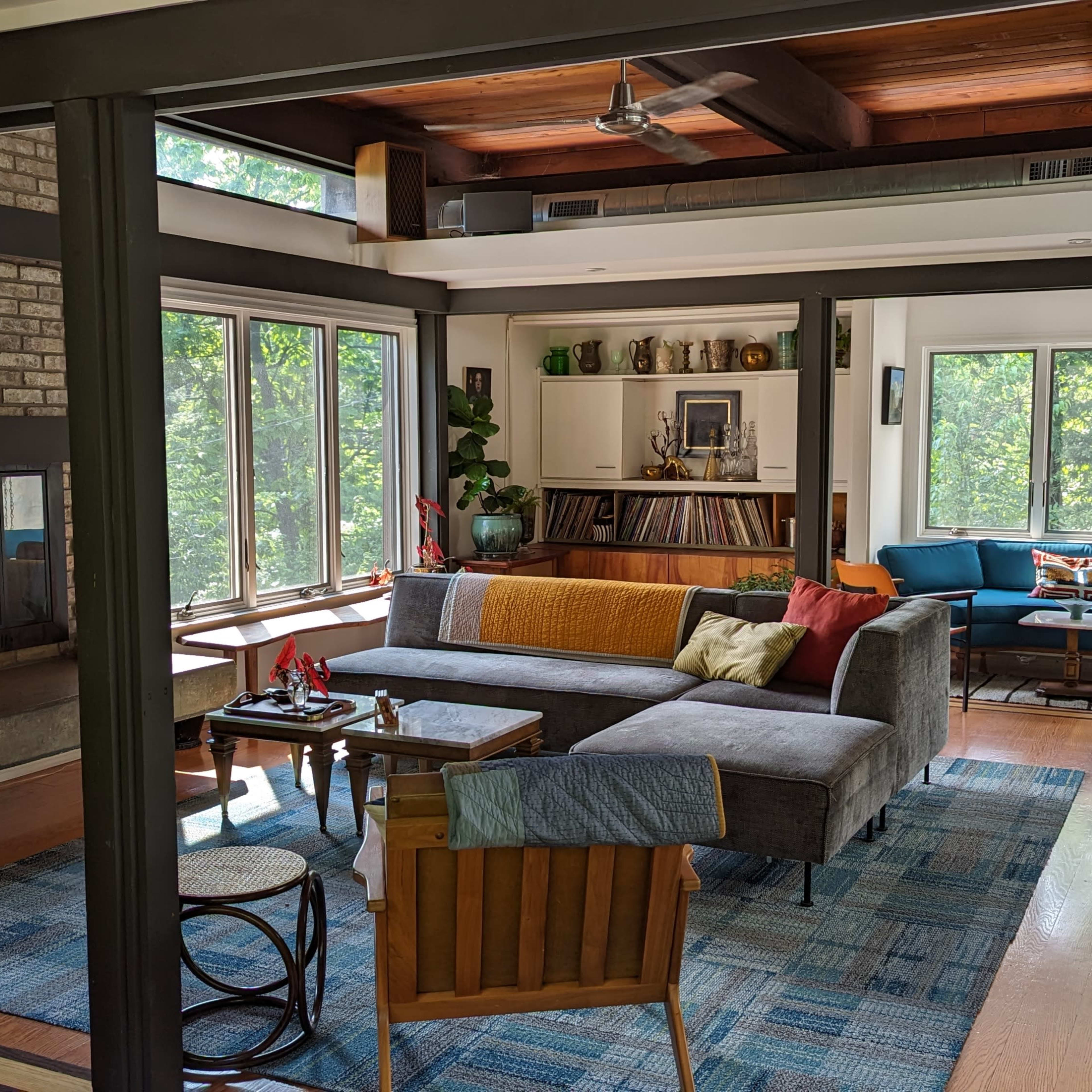 A modern living room featuring a gray sectional sofa, a brick fireplace, large windows, and a mix of wooden and upholstered furniture.