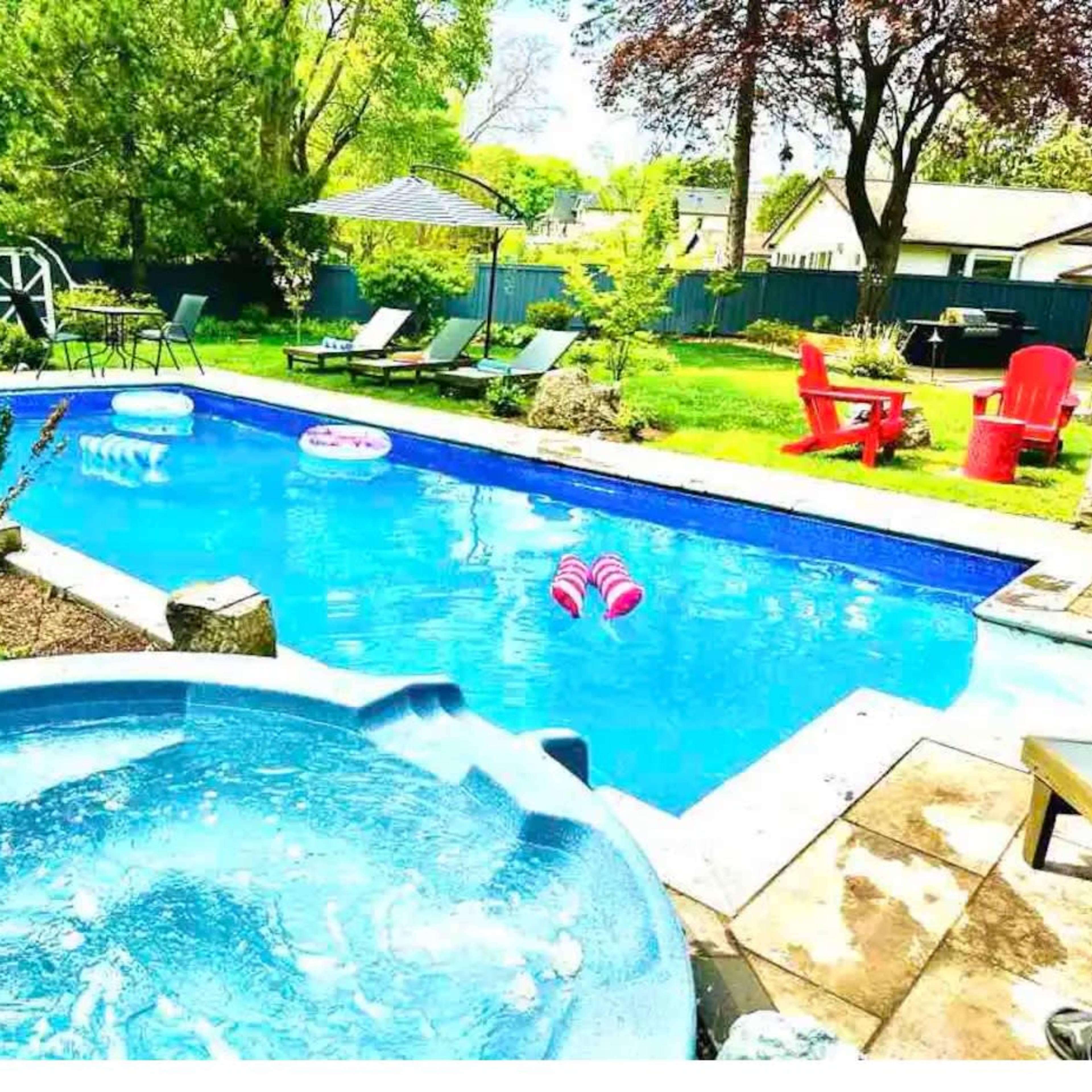 A backyard pool area with a hot tub, lounge chairs, and colorful pool floats surrounded by greenery.
