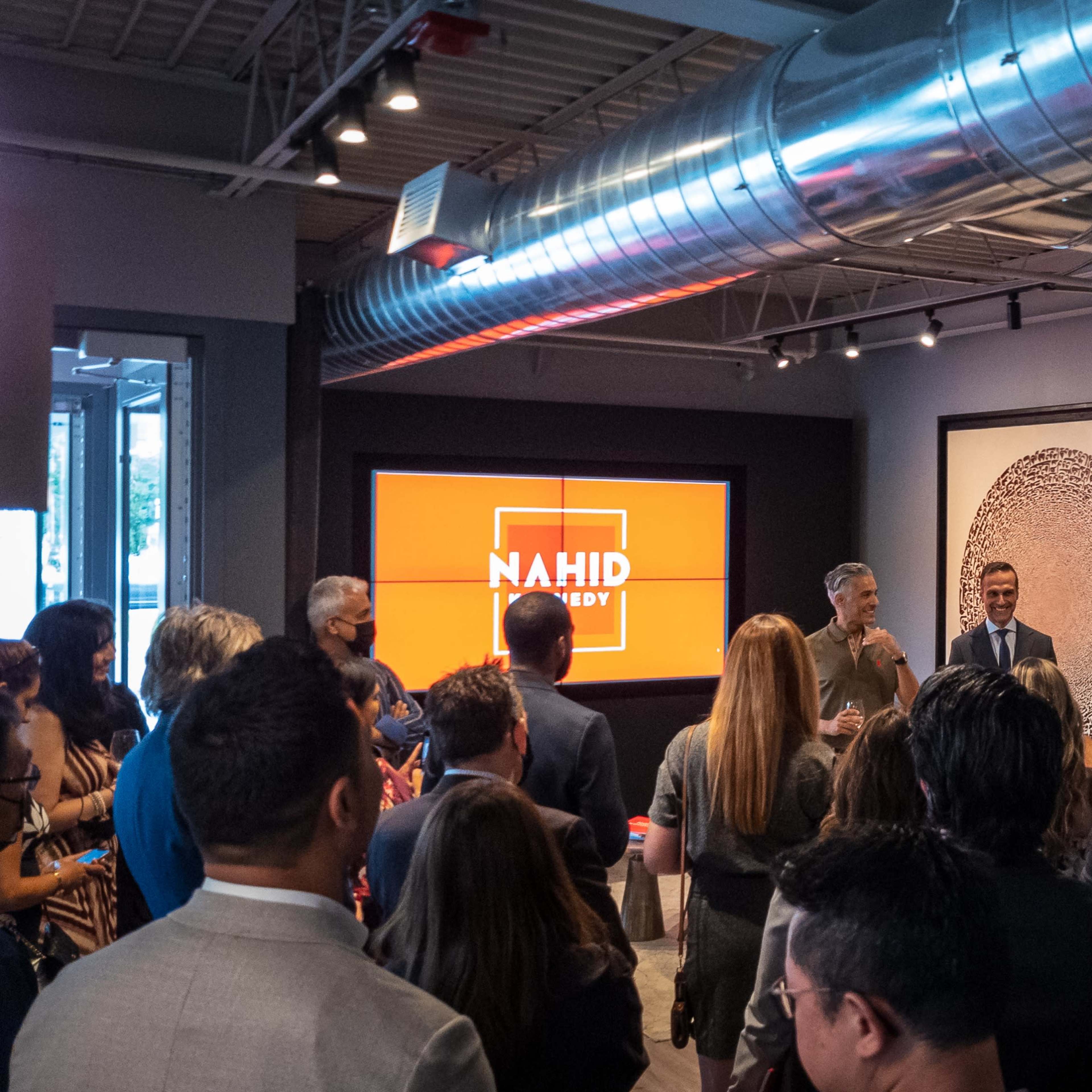 A group of people stands in a contemporary event space, while two speakers address the audience in front of a large screen displaying the name "NAHID" above a circular artwork.
