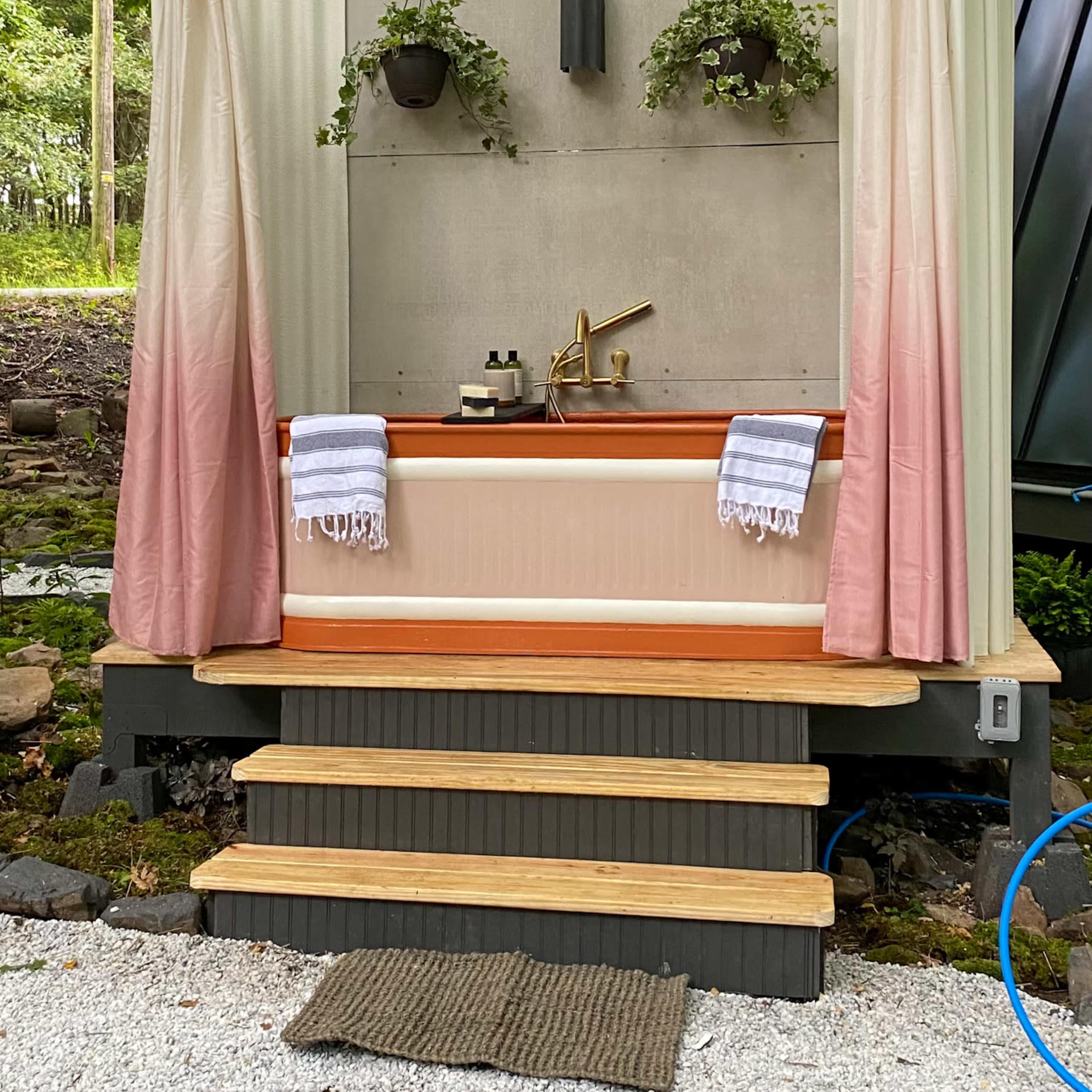 An outdoor bathing area with a wooden tub, two towels hanging, and curtains on either side, all set against a backdrop of greenery and stairs leading up to it.