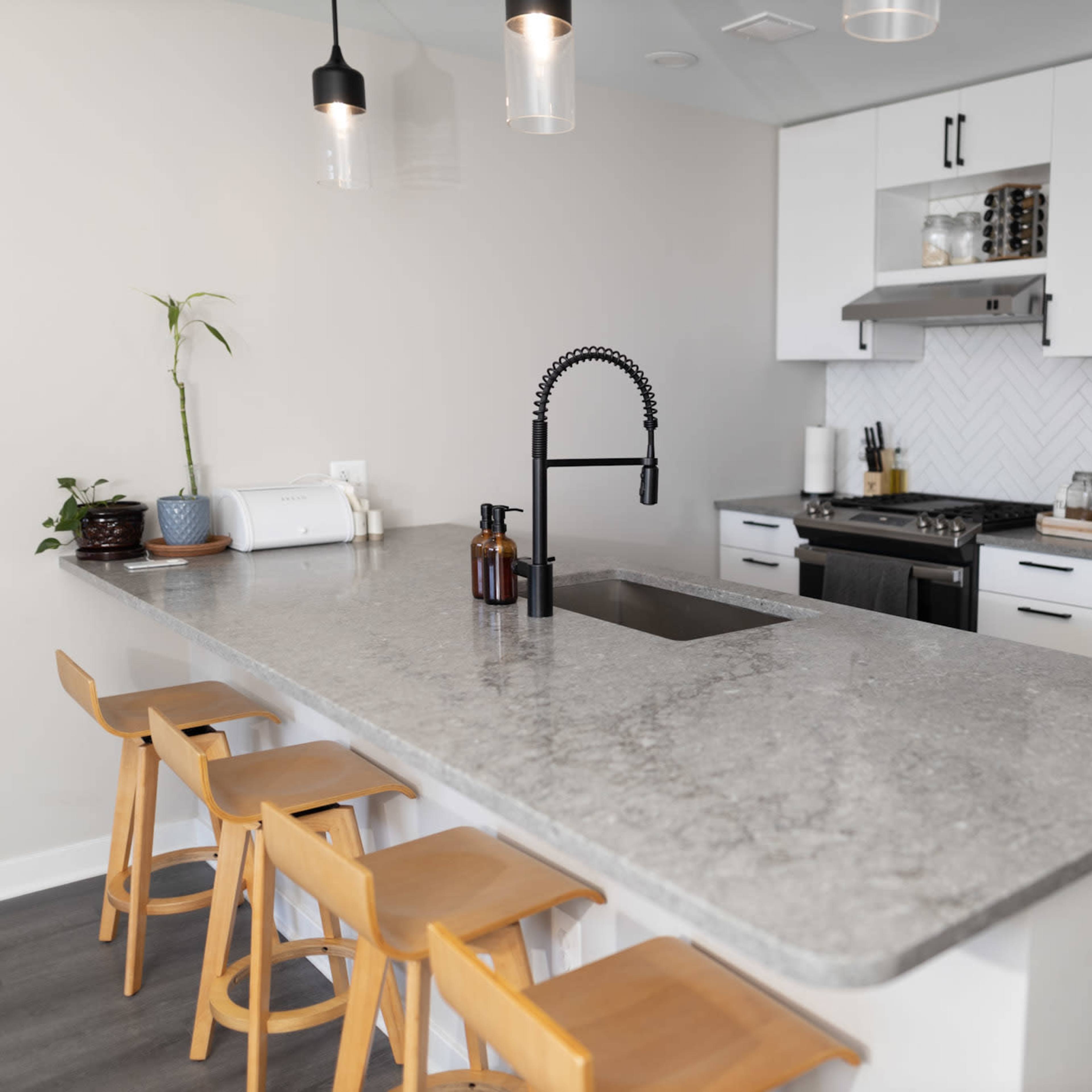 A modern kitchen featuring a light-colored countertop, stainless steel appliances, and four wooden stools at a breakfast bar.