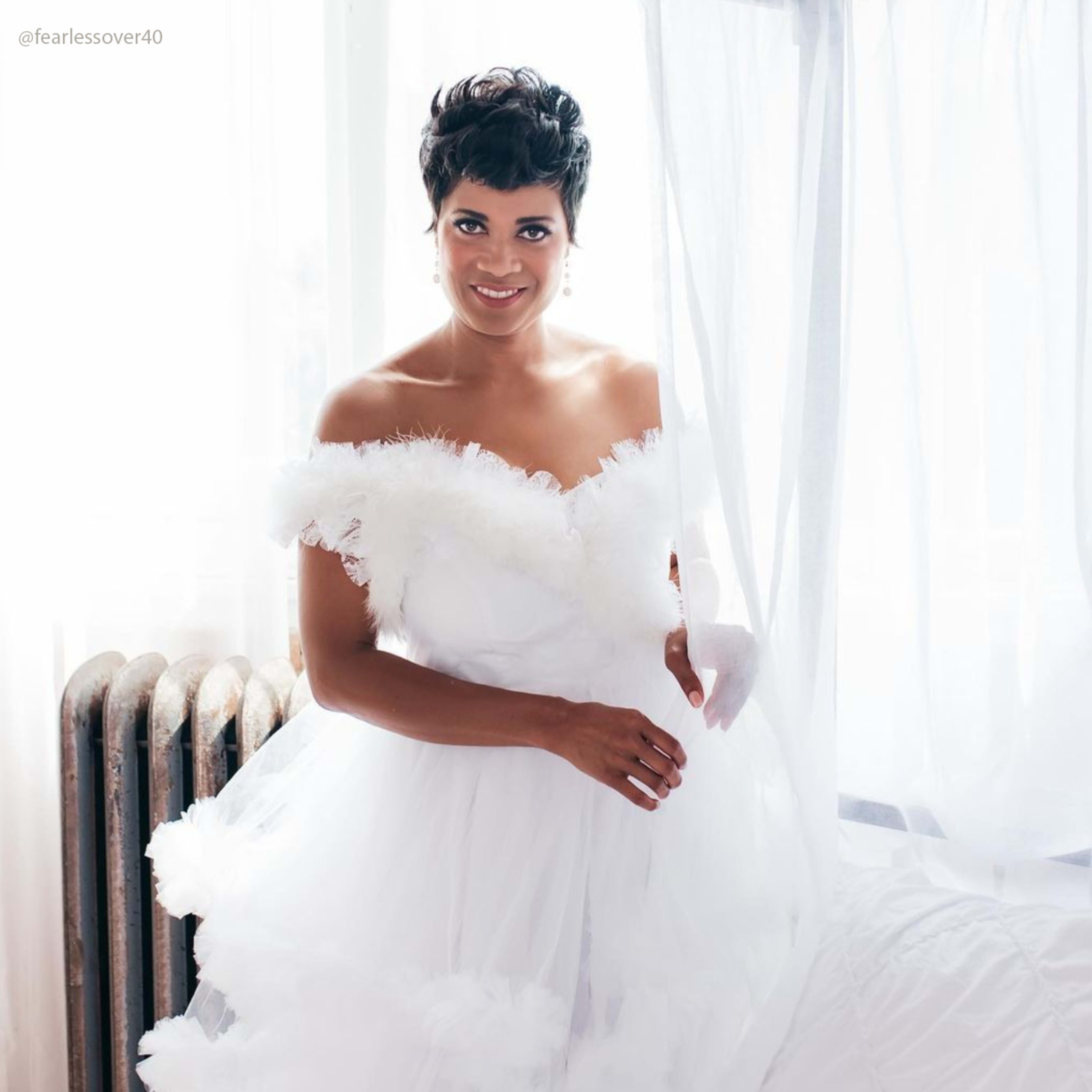 A woman in an off-the-shoulder white dress stands by a window with sheer curtains.