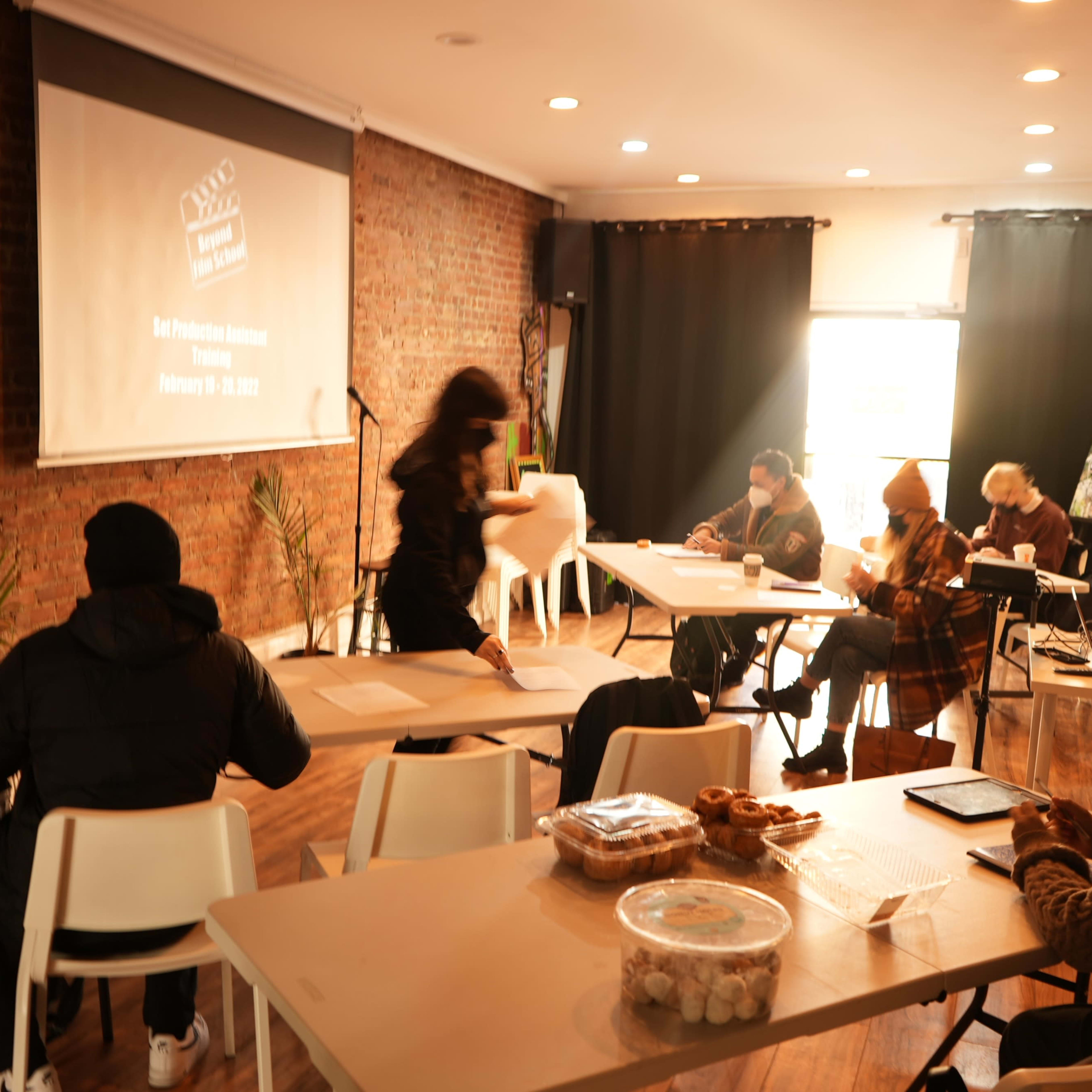 A group of people sits at tables in a well-lit room with exposed brick walls, watching a presentation while some work on laptops.