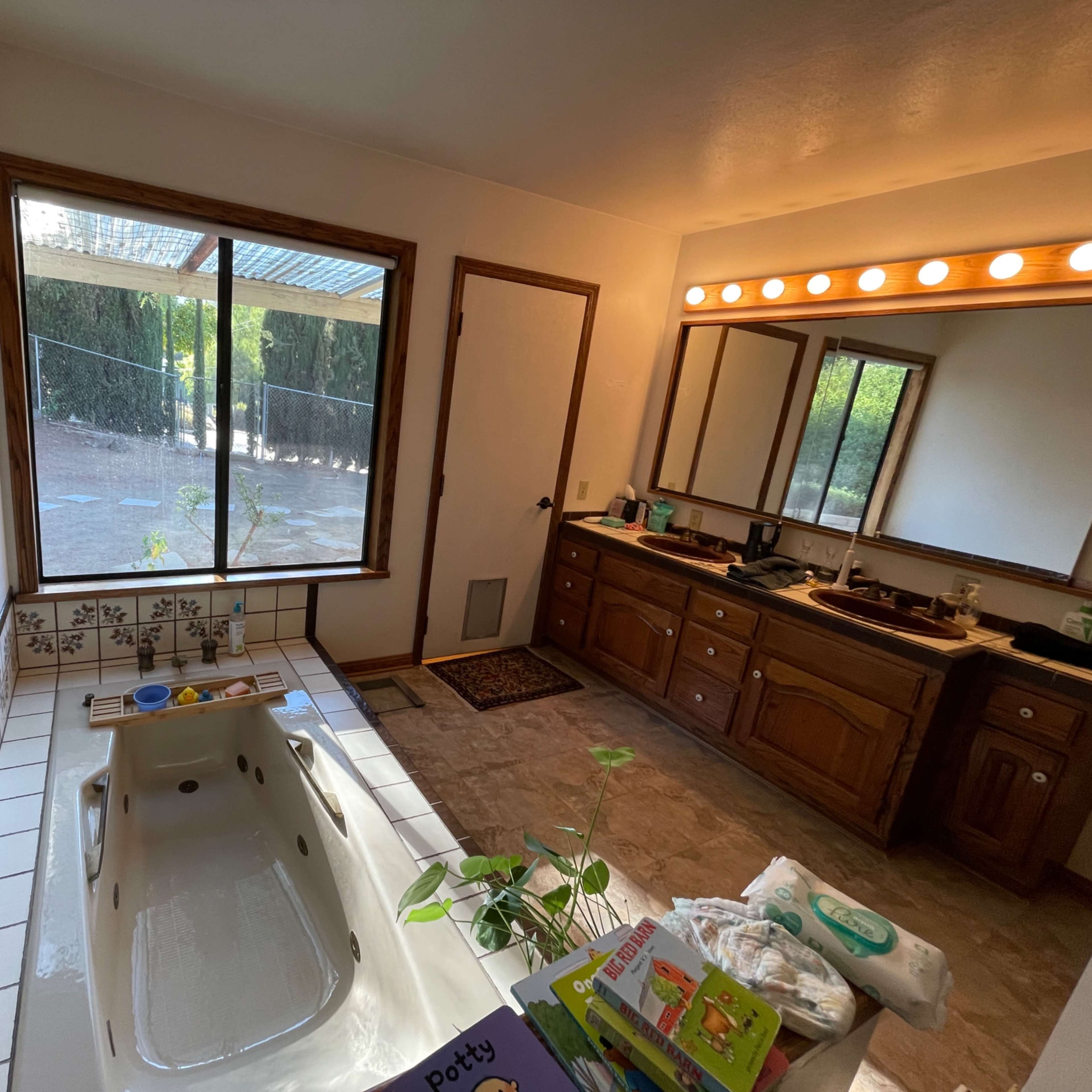 A bathroom with a jetted tub, twin sinks, a large window overlooking a yard, and various items on the countertop and floor.