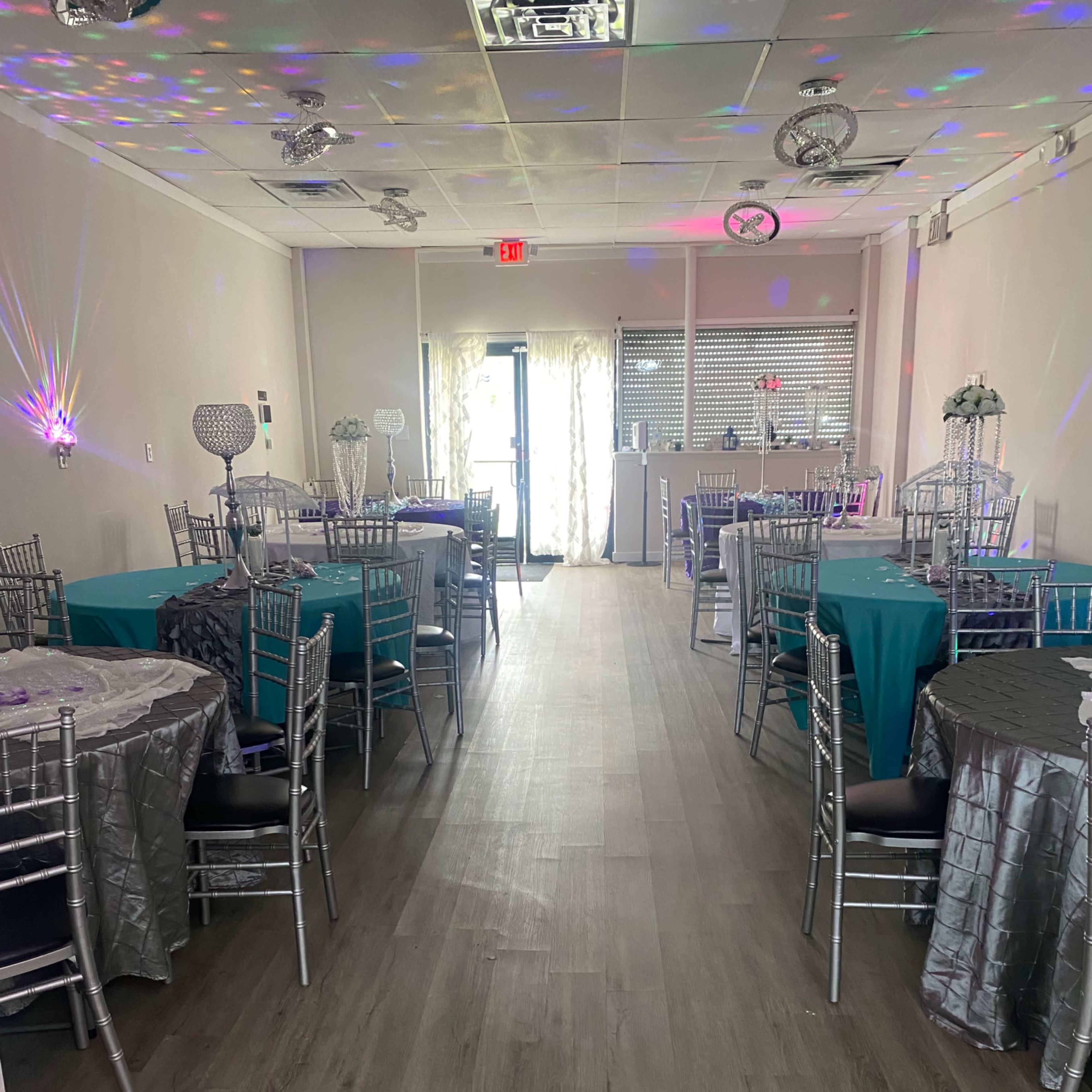 An event space with multiple tables dressed in teal and silver linens arranged neatly, leading to a doorway at the far end.