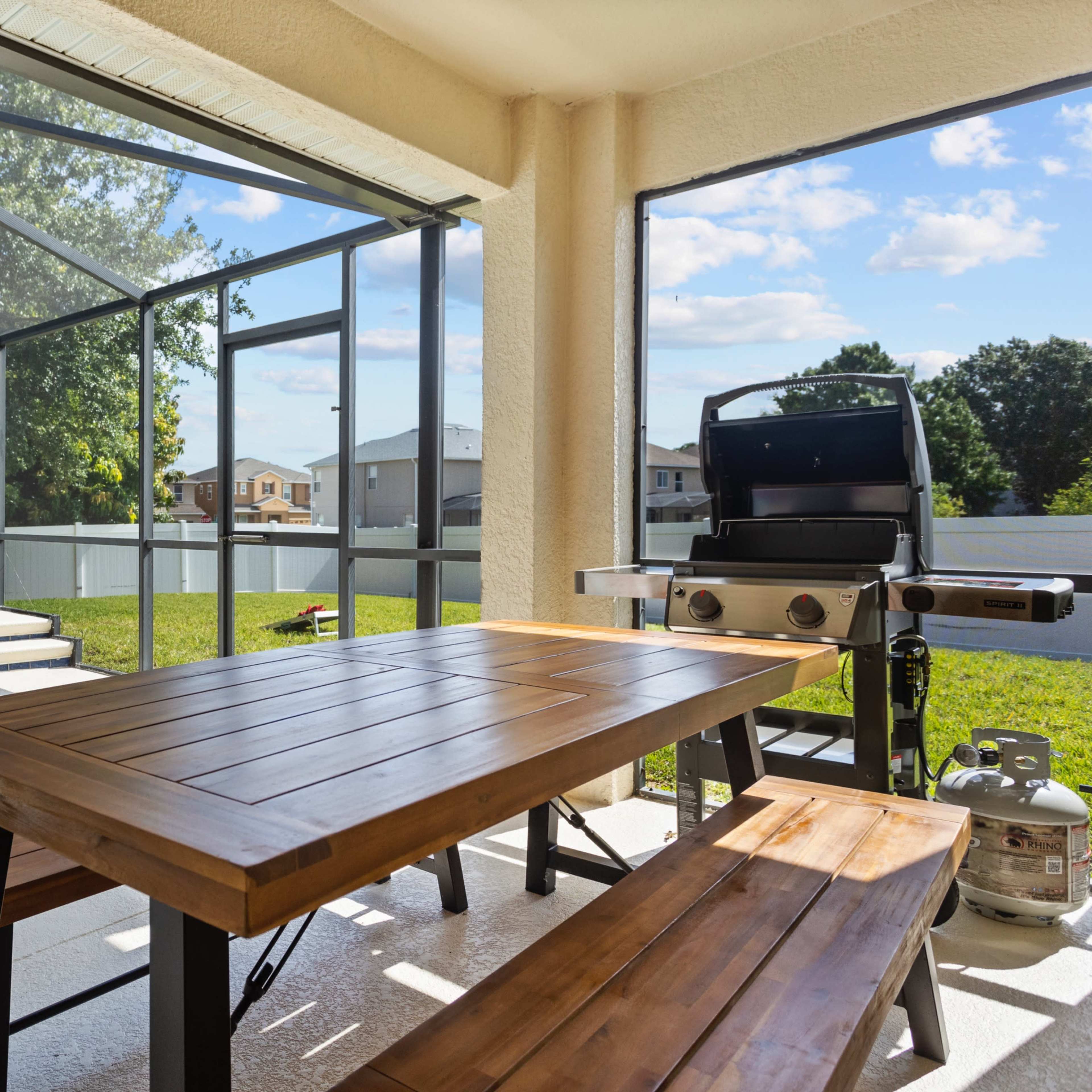 A covered patio features a wooden dining table beside a grill, overlooking a pool and backyard enclosed by a screen.