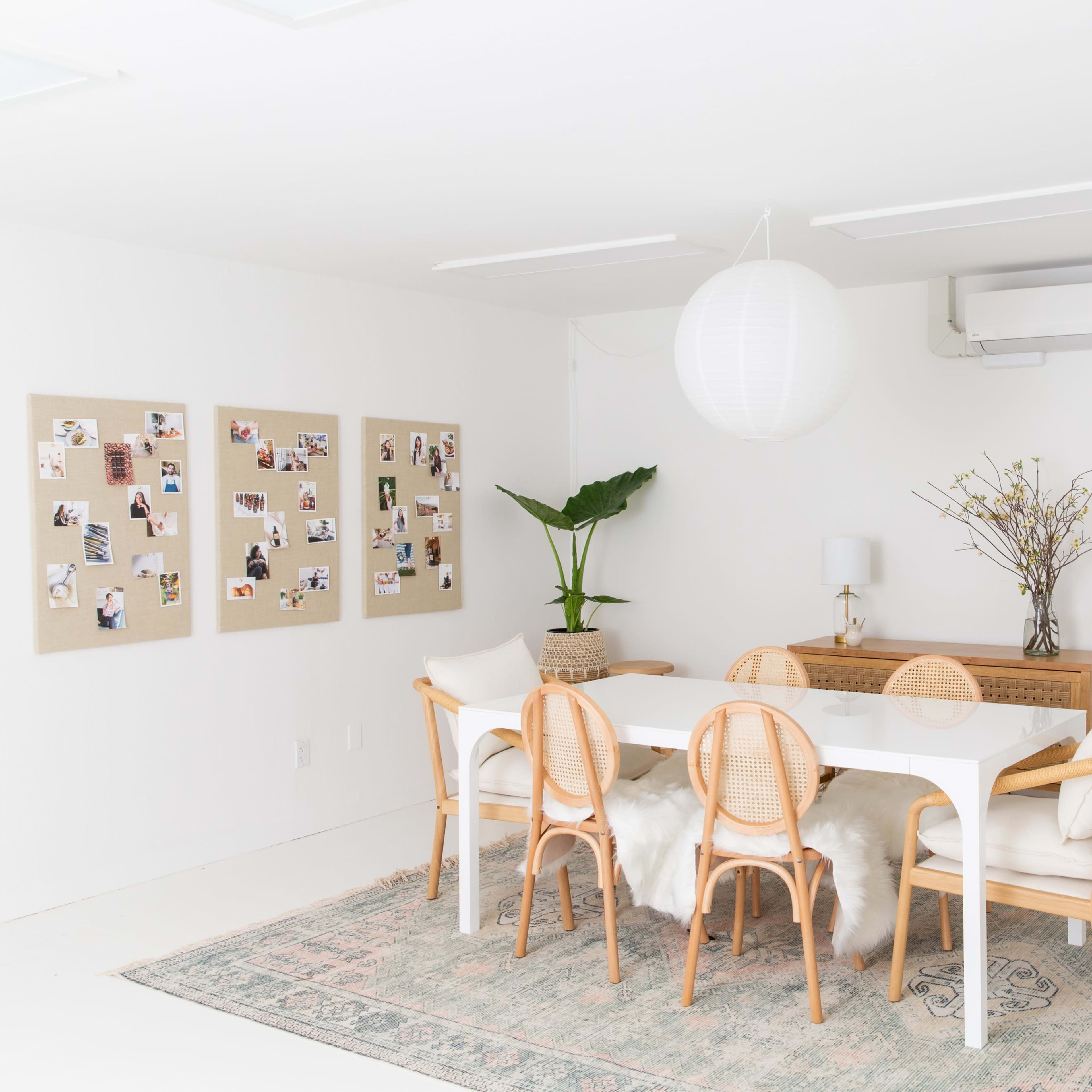 A bright, modern dining room features a white table surrounded by wooden chairs, a gallery wall of photos, and a large mirror.