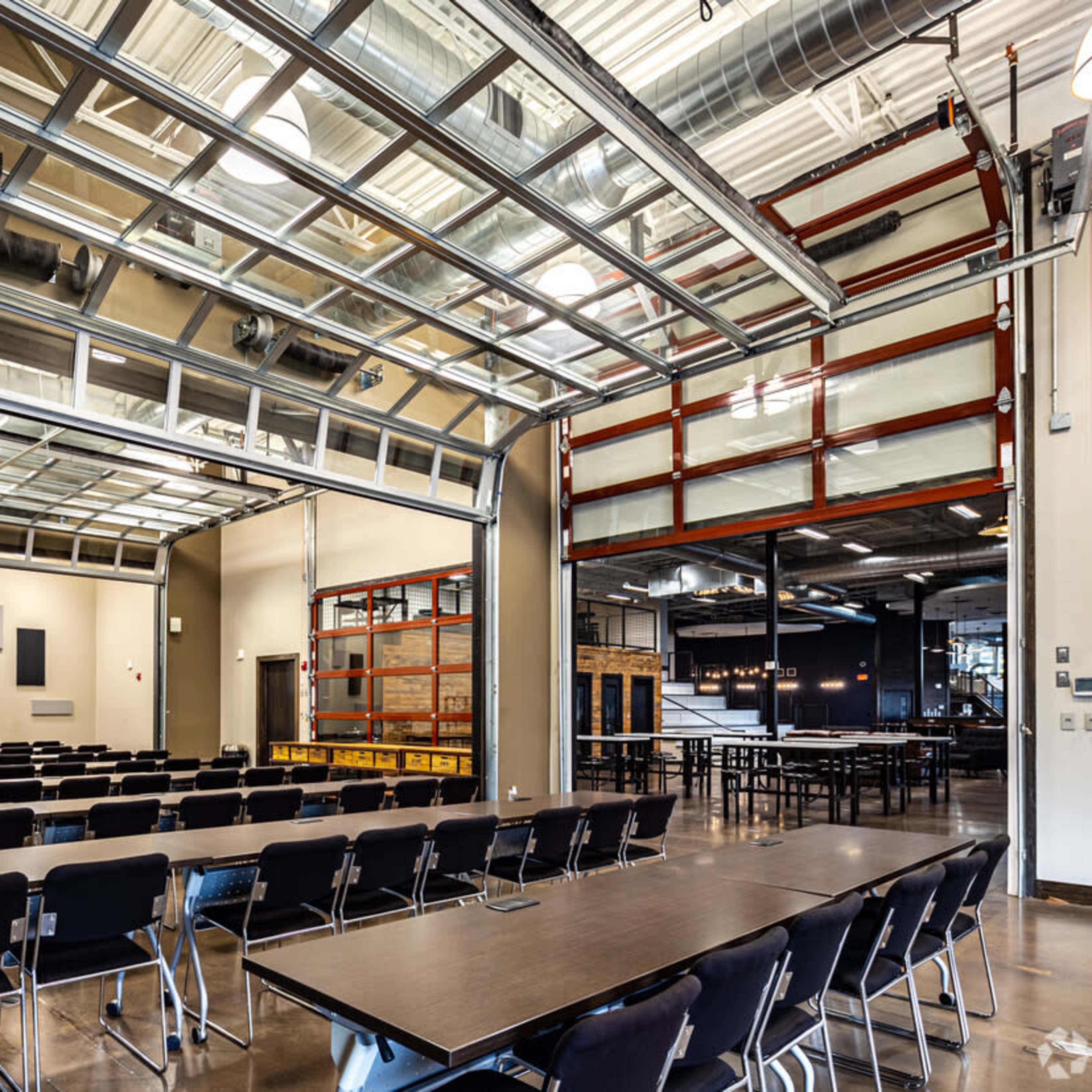 A modern conference room with rows of black chairs facing a projector screen, featuring large glass doors that open to an adjoining space.