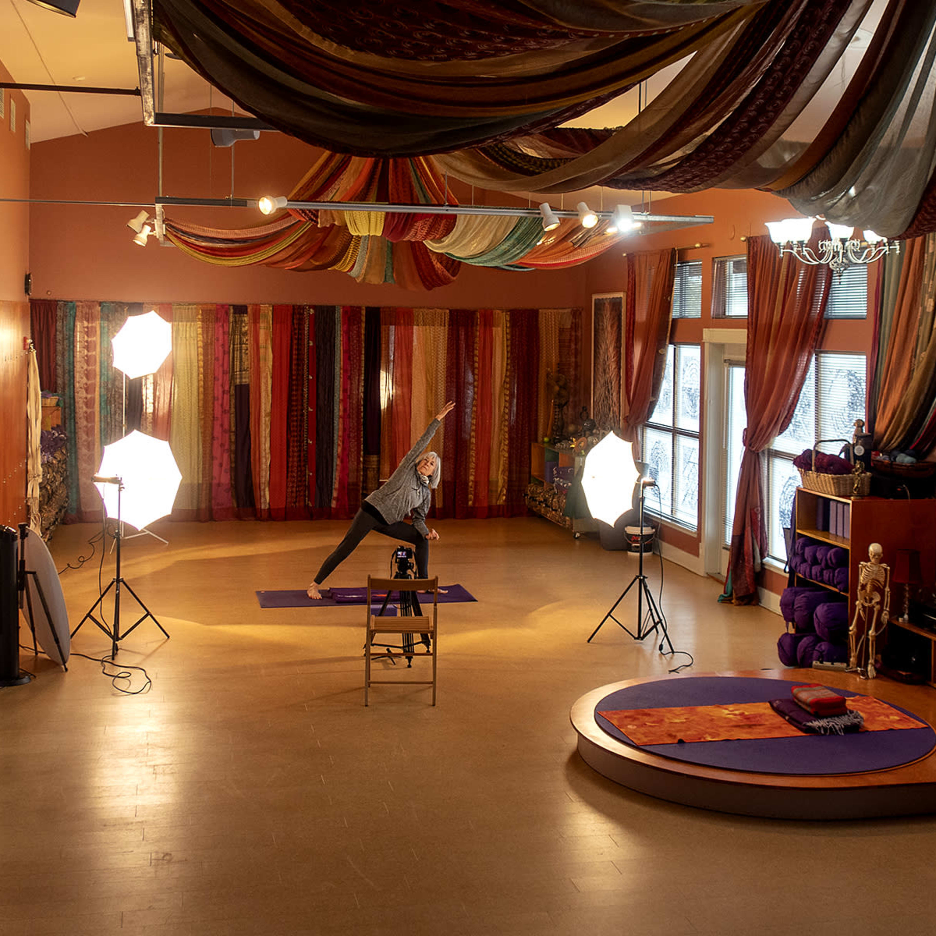 A yoga studio features a person practicing a pose on a mat, surrounded by colorful fabric drapes and studio lighting equipment.