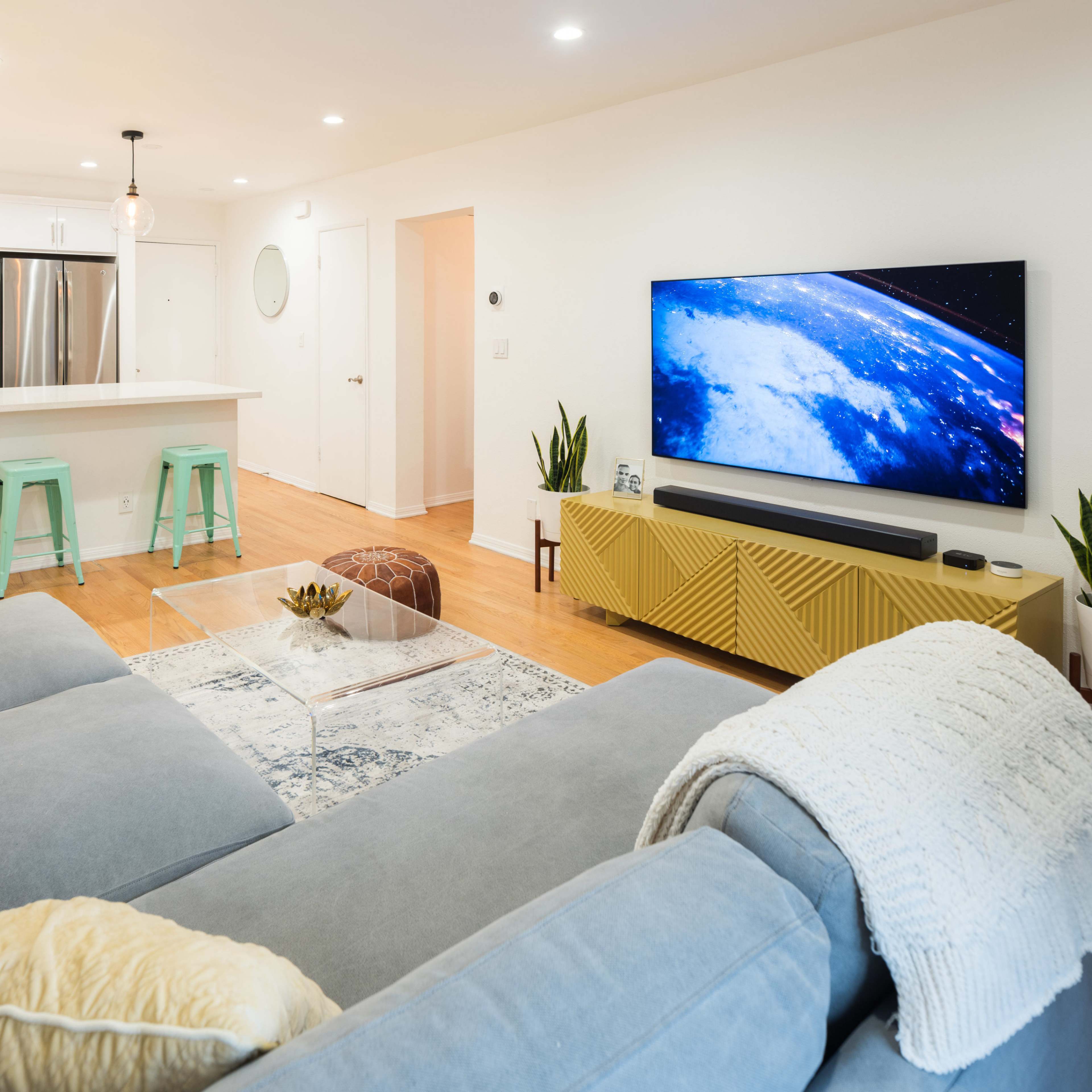 A modern living room with a gray sectional sofa, a glass coffee table, a large television mounted on a yellow media console, and a bright kitchen area in the background.