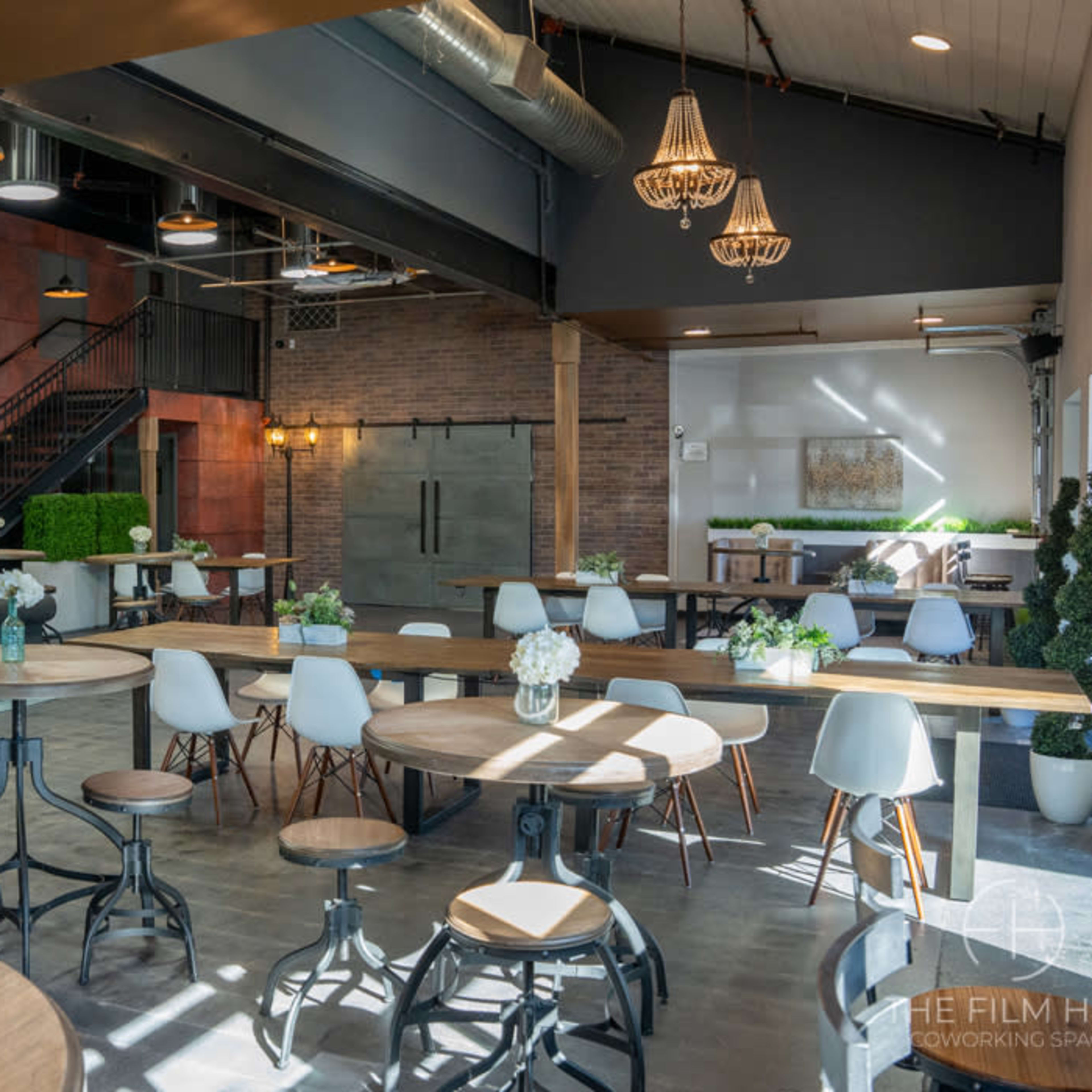 A modern coworking space with wooden tables, green plants, and a staircase in a well-lit interior featuring brick walls and industrial design elements.