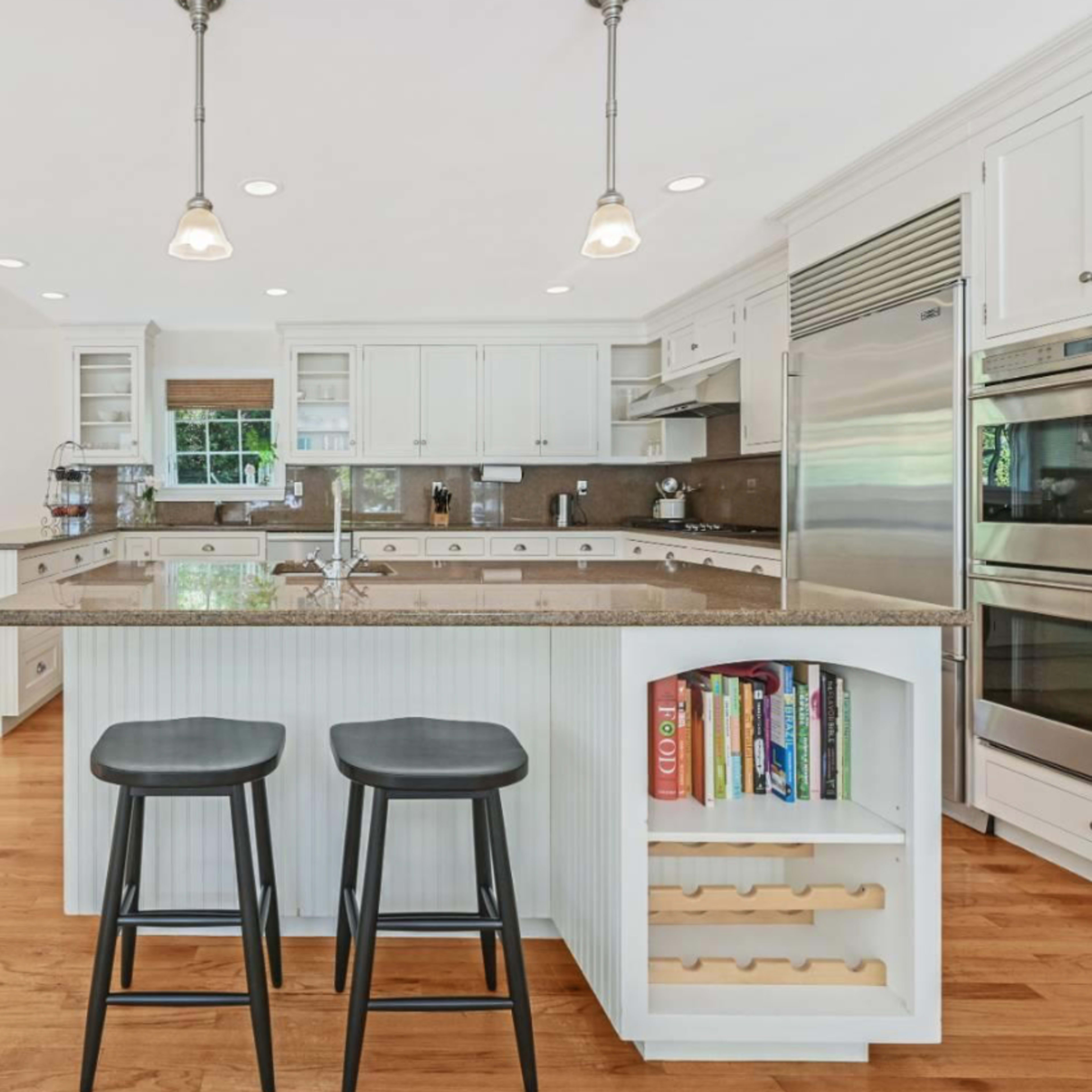 A spacious kitchen features an island with two black stools, stainless steel appliances, and a dining area with wooden chairs.