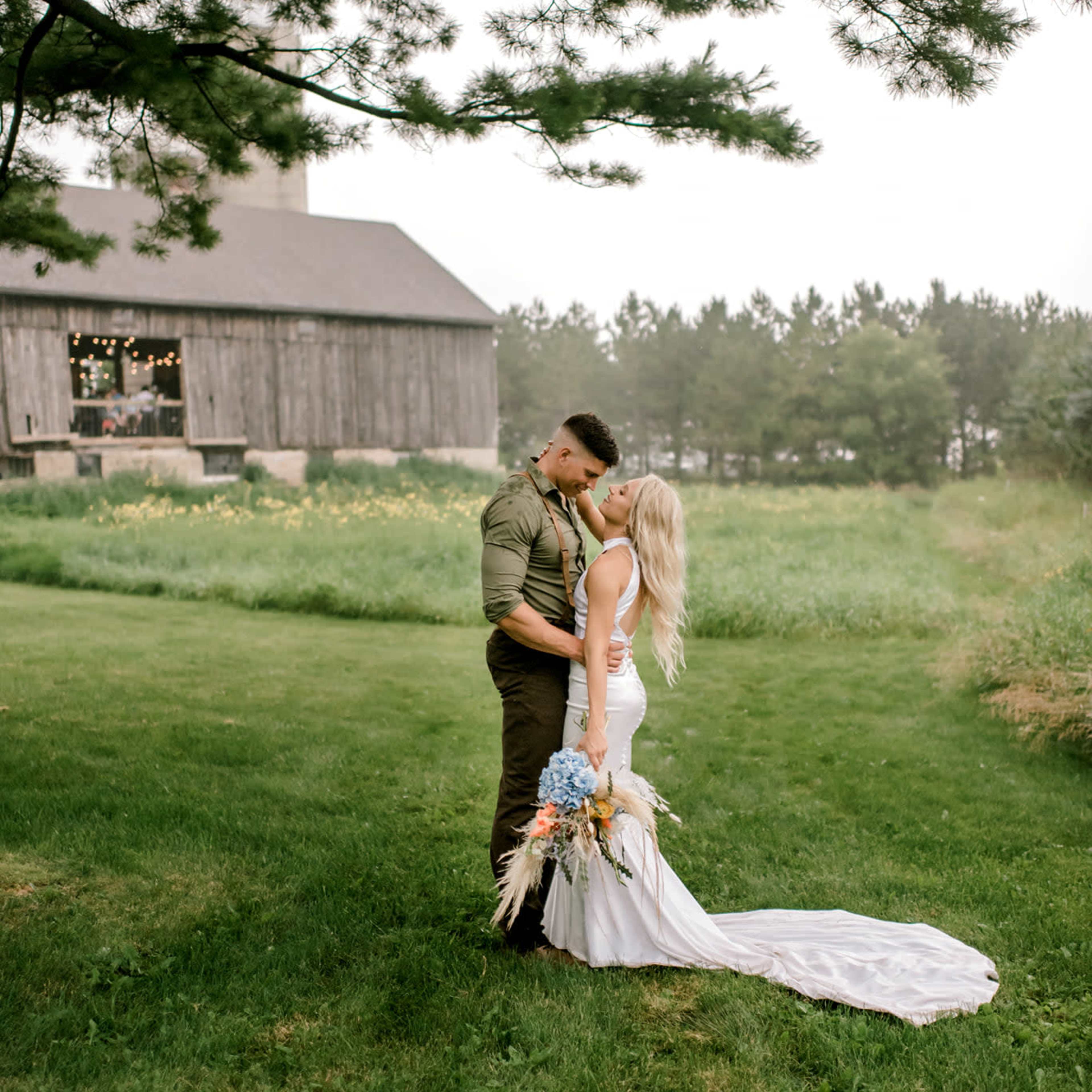 A couple stands in an open field, embracing each other with a rustic barn and trees in the background.