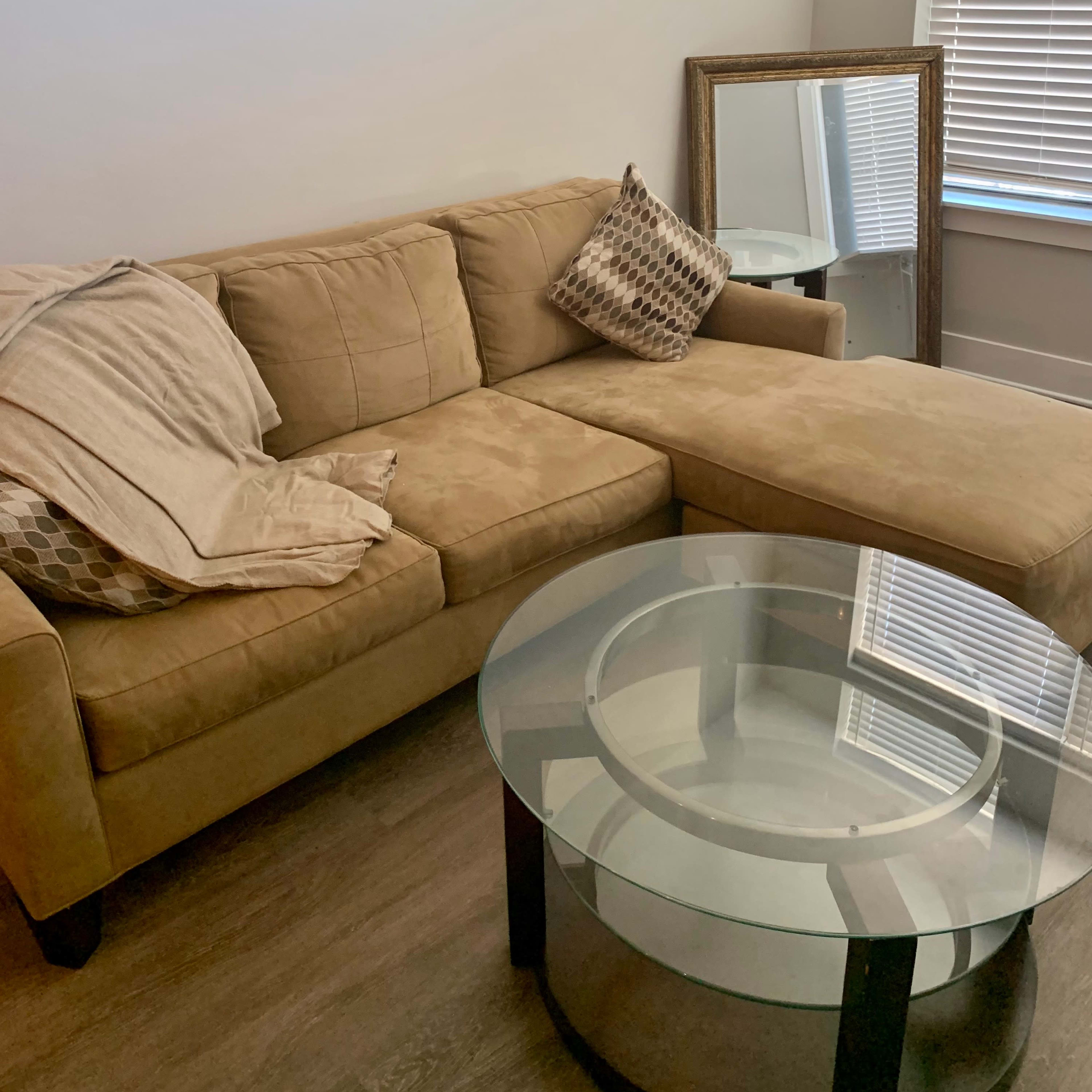 A tan sectional sofa with a decorative pillow is positioned next to a round glass coffee table and a mirror, under a window with blinds.