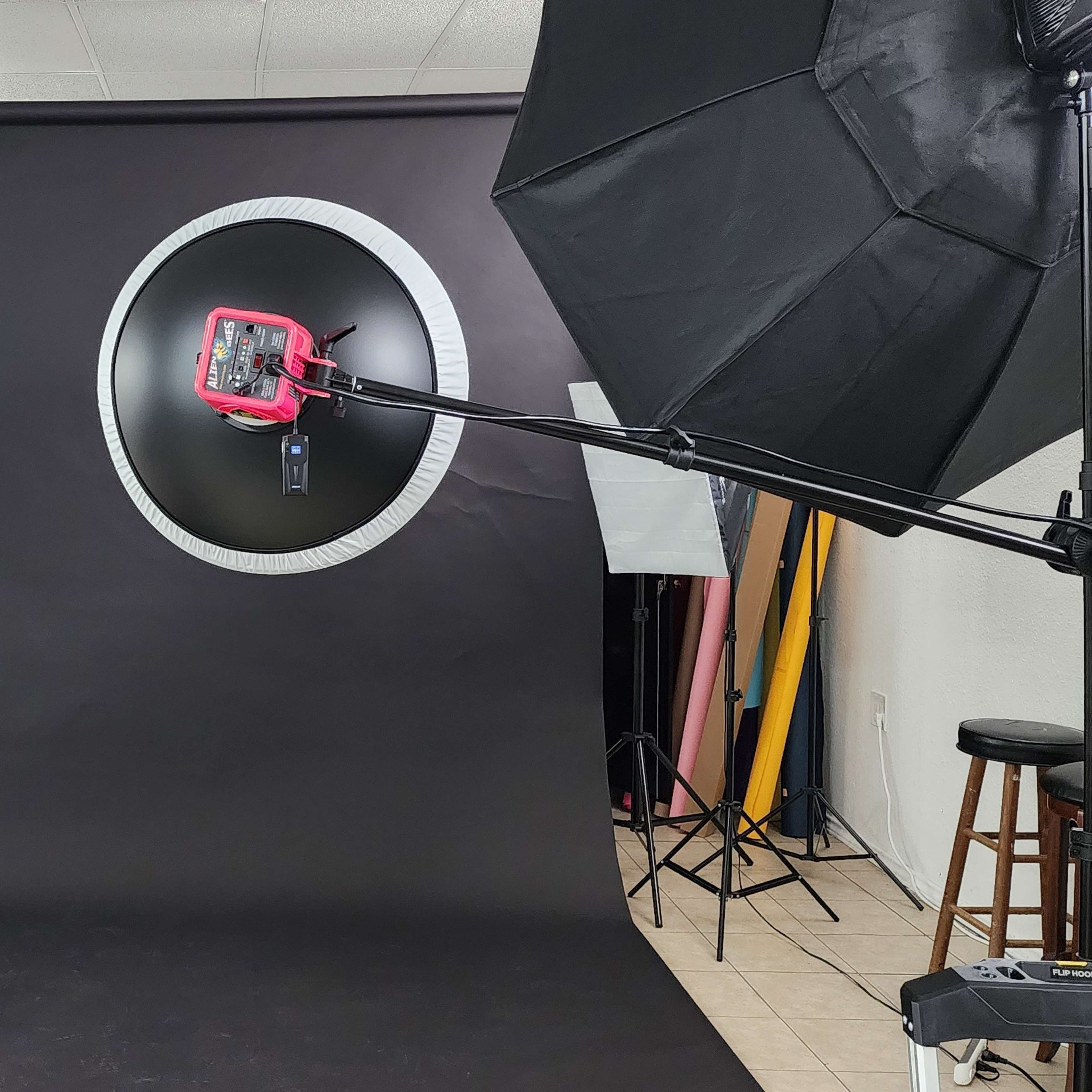 A photography studio setup featuring a black backdrop, two softbox lights, a ring light on a stand, and a variety of colorful drapes hanging on the side.