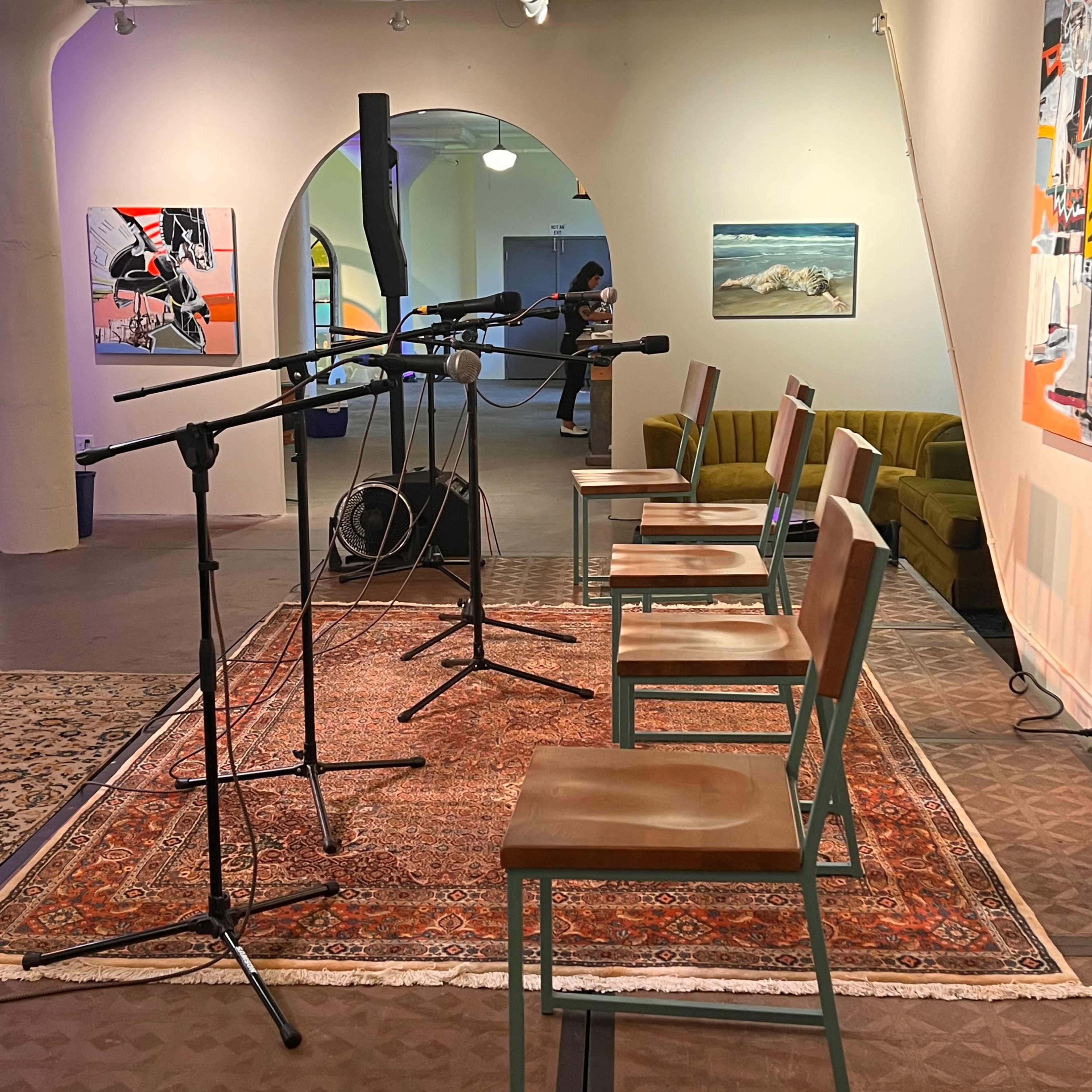A small stage setup features five chairs arranged in a semicircle around multiple microphones, with artwork and a green sofa visible in the background.