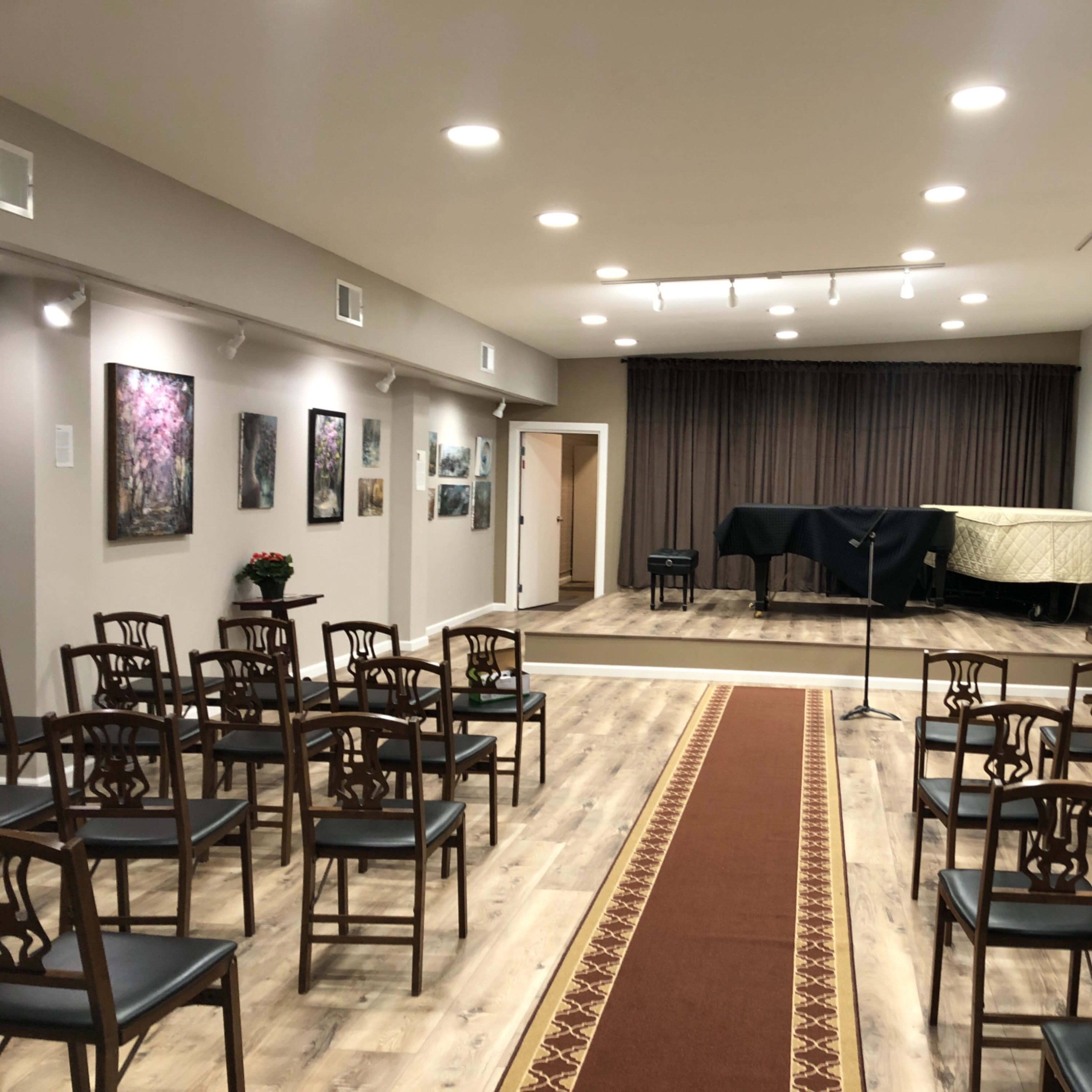 A music performance space with rows of wooden chairs facing a stage covered with a black cloth and a piano, surrounded by framed artwork on the walls.