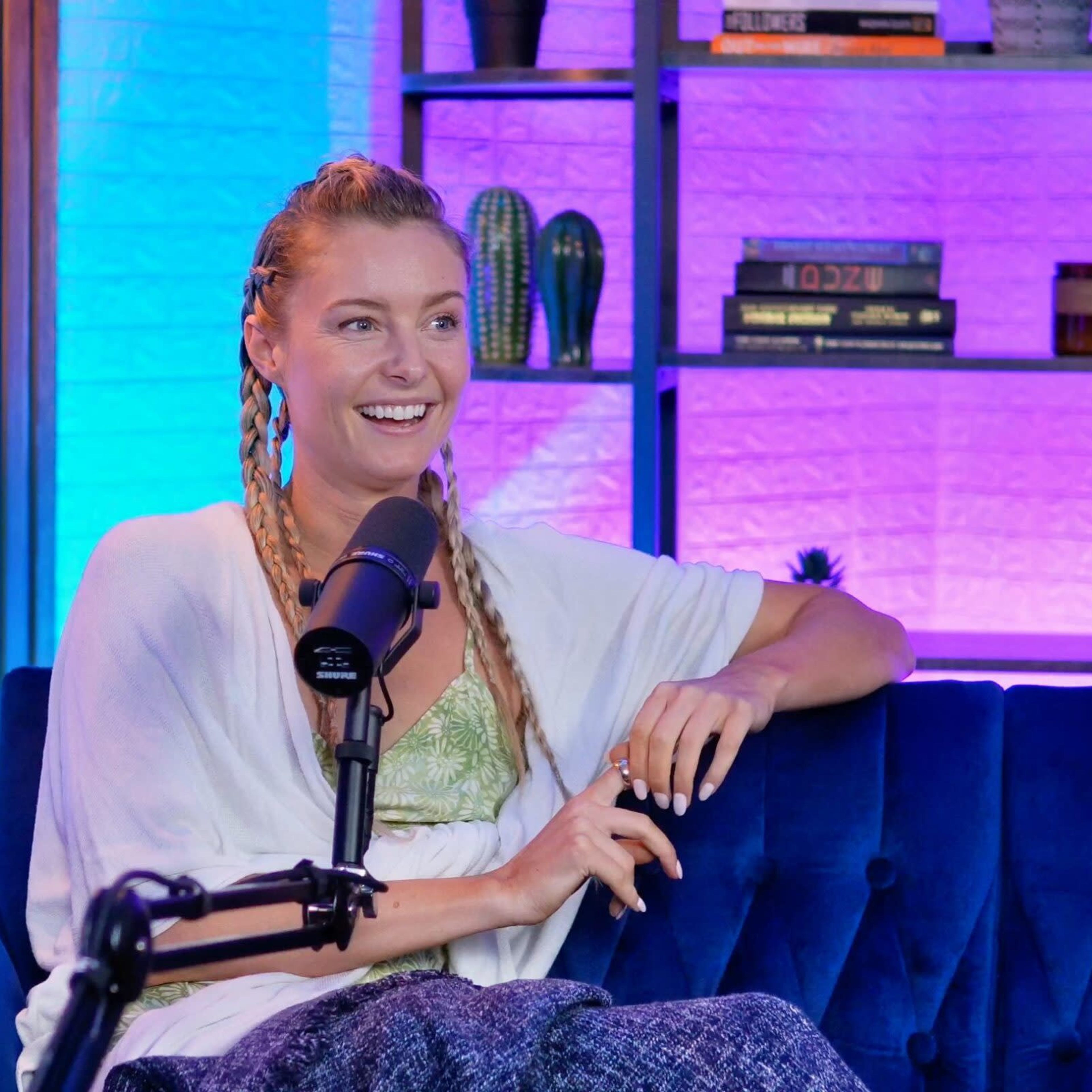 A woman with braided hair is sitting on a blue couch, smiling while speaking into a microphone in a brightly lit studio with colorful background lighting and bookshelves.