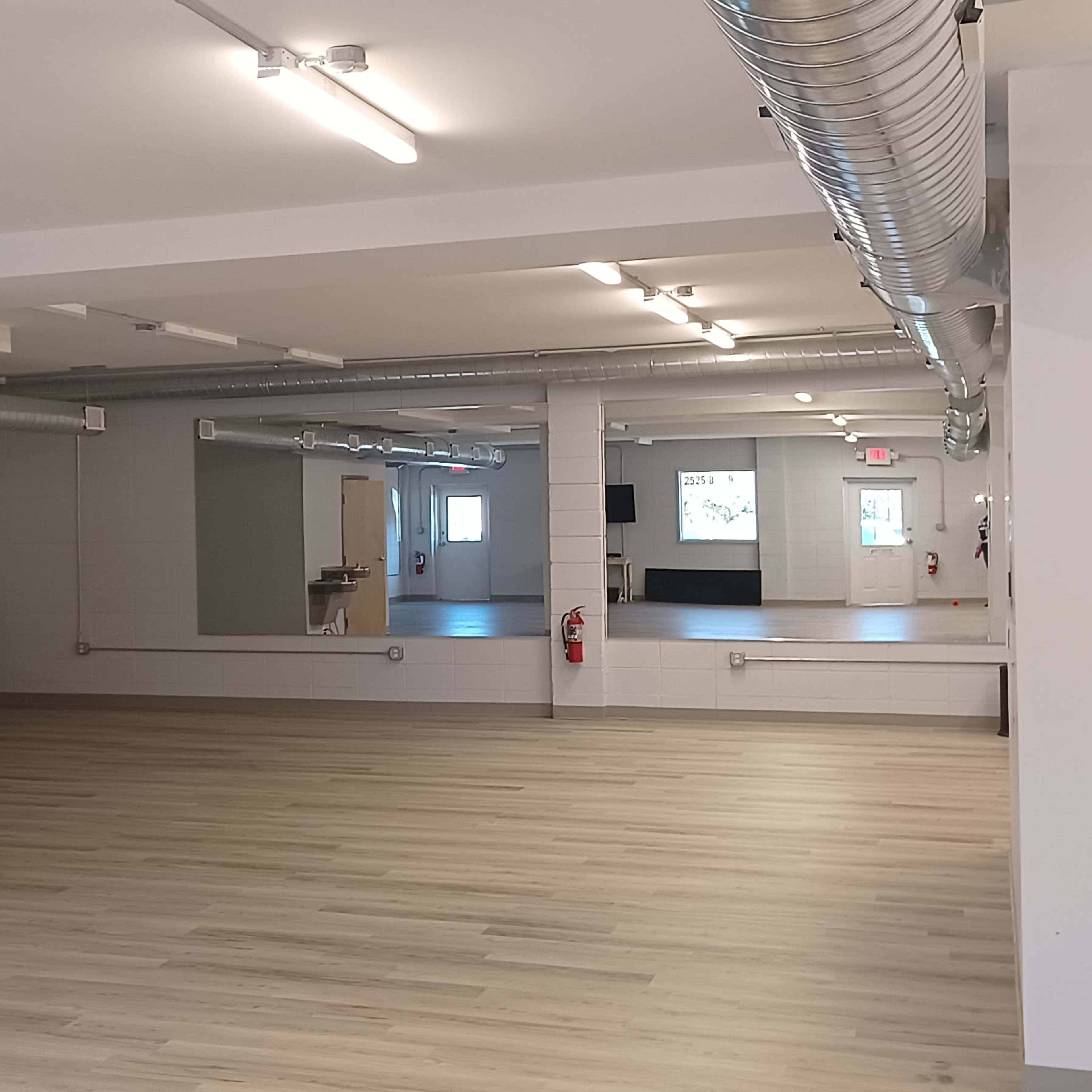 A spacious, empty room with wooden flooring, industrial lighting, and a large mirror on one wall.
